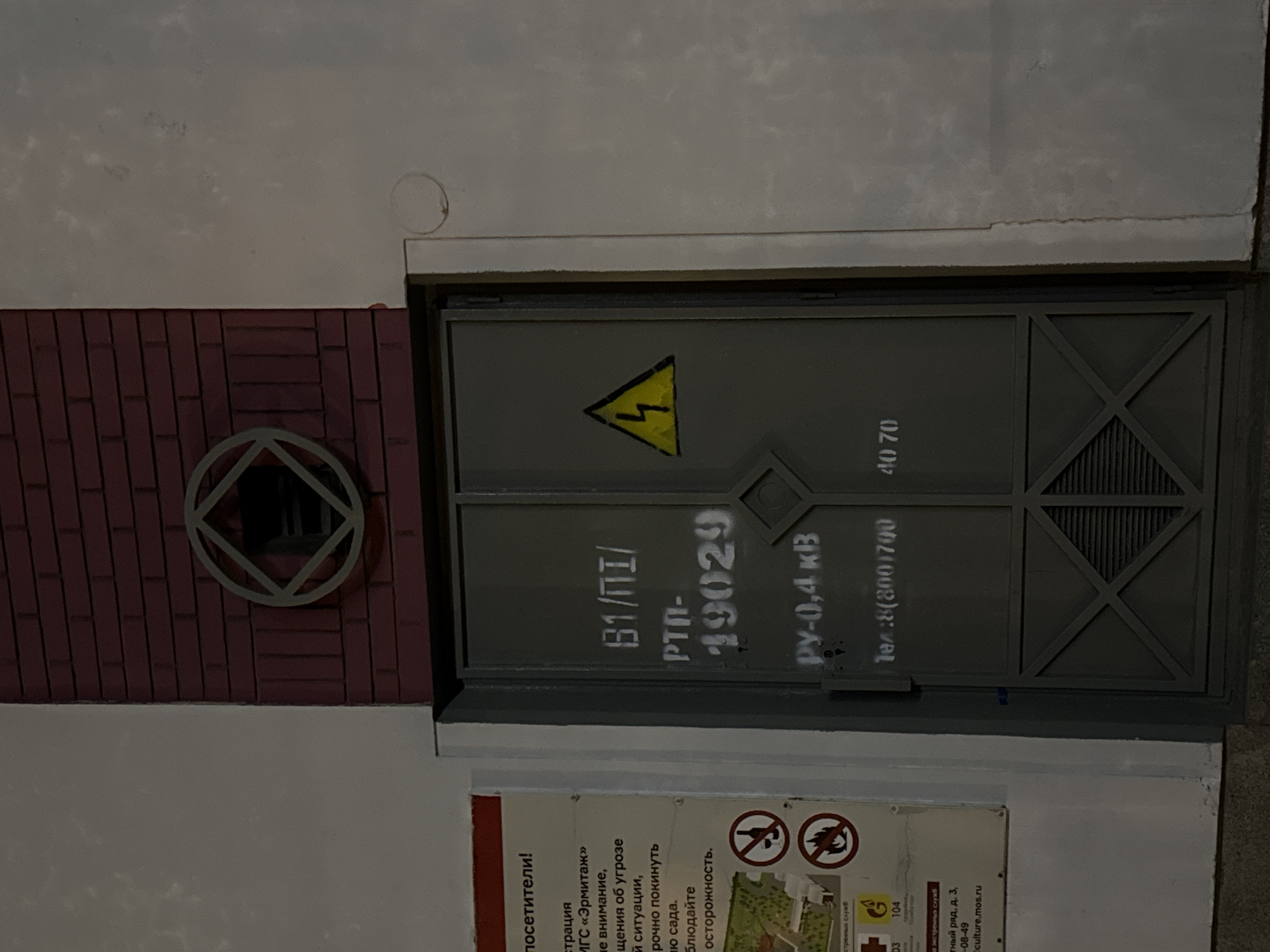 The image shows a gray electrical panel or utility box with a yellow lightning bolt symbol on its door, indicating a high voltage area. The panel door features stencil-like lettering that reads "B1/ΠΙ/ РТП-19029 РУ-0,4 кВ" and "Тел: 8(800)700 4070". The box is set into a wall next to a small, decorative window with a geometric design, framed by red brickwork. Below the box, a sign with visitor information is visible, featuring Russian text and pictograms, including symbols for no open flames and no swimming. The wall around the box is a light gray or white color. The image appears to be taken in a city setting, possibly a public park or garden.