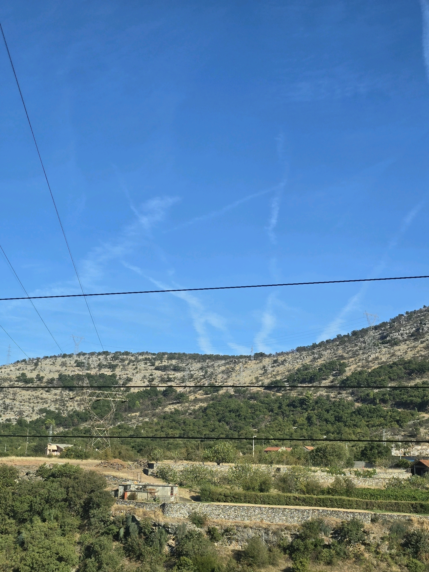 The image shows a hilly landscape with scattered houses and vegetation under a bright blue sky with contrails. Several power lines are visible in the foreground and middle ground, stretching across the frame. The terrain is dry and rocky in some areas, with patches of green trees and shrubs. There are no people or animals visible in the image. The scene appears to be a rural area, possibly on the outskirts of a city. The specific location in Podgorica, Montenegro is not discernible from the image itself, but the landscape is consistent with areas around Podgorica, which is situated in a valley surrounded by mountains.