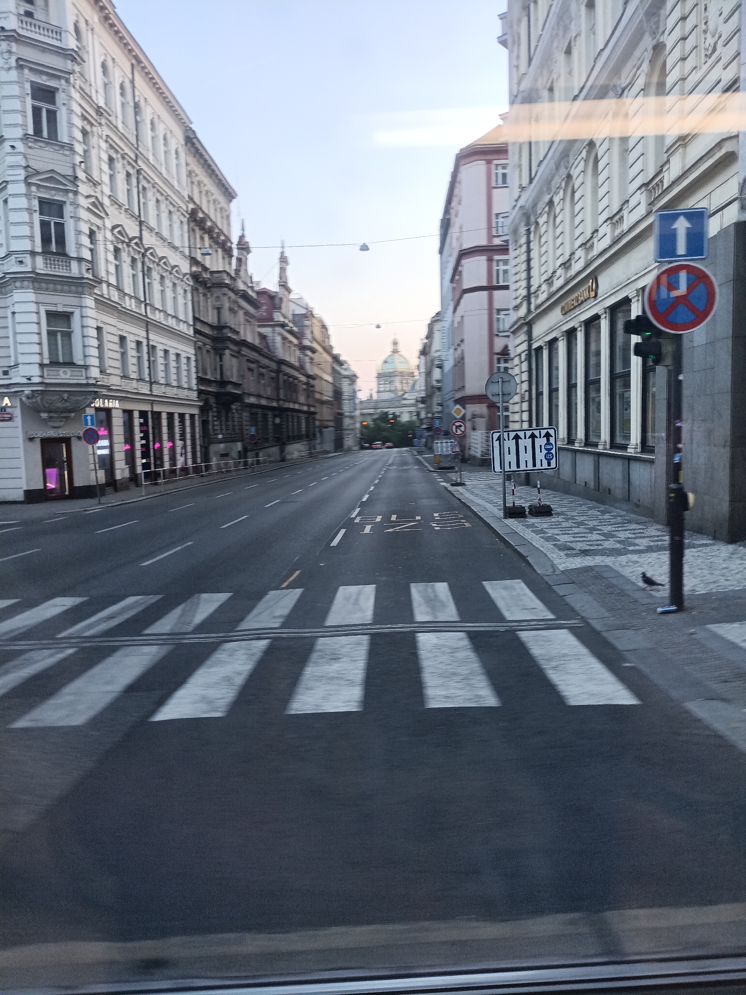 The image shows a street scene in Prague, Czechia. The street is lined with multi-story buildings on both sides, with architectural styles suggesting an urban European setting. A pedestrian crossing is prominently visible in the foreground. Traffic signs are present, including one indicating a one-way street with an upward arrow and another with a red circle and a diagonal bar over a blue square with a white arrow, signifying no stopping or parking. A traffic light is also visible, appearing to be green. In the distance, a domed structure, possibly a church or historical building, can be seen. The sky is overcast, suggesting early morning or late afternoon. There is a solitary bird on the sidewalk. The perspective suggests the image was taken from a moving vehicle, possibly a tram, due to the presence of parallel tracks on the road. The overall impression is of a quiet city street, likely with moderate traffic during peak hours.