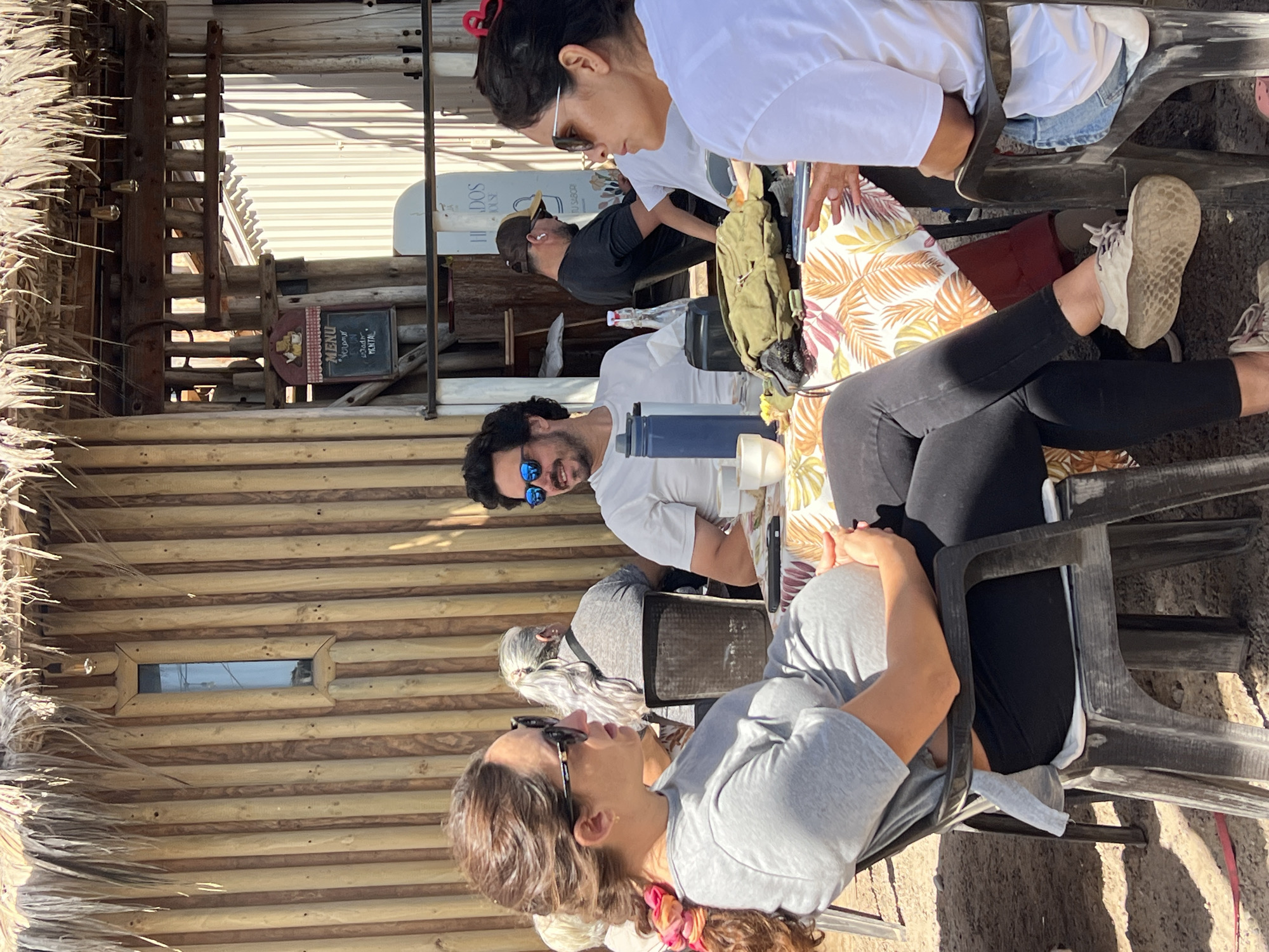 The image depicts a group of people seated at an outdoor establishment. There are at least four individuals visible at the table. They are engaged in conversation. One man is wearing blue sunglasses and smiling. Another woman is wearing sunglasses and a white shirt, her hair pulled back with a red hair tie. The location features a wooden structure with a thatched roof, possibly a restaurant or bar. A sign with a menu is also visible. The scene is likely located in Arica, Chile, given the request to include the city.