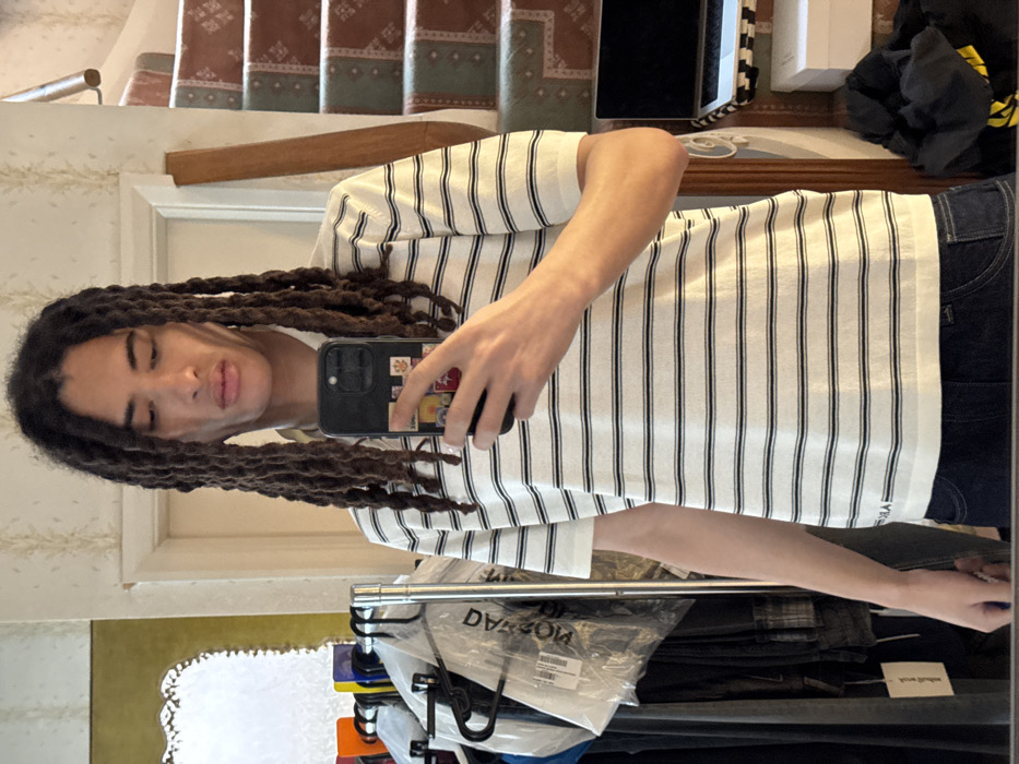 The image displays a young individual with long, dark dreadlocks, positioned in an indoor residential setting, likely in London, United Kingdom. The person wears a short-sleeved white polo shirt with thin horizontal dark stripes and dark blue jeans, actively taking a mirror selfie. A black smartphone, featuring a case adorned with multiple colorful stickers, is held in the subject's right hand.
The background on the left side of the image reveals a clothing rack laden with various garments, some encased in clear plastic bags; one garment clearly displays the reversed text "balenciaga." Above the rack, a partially visible ornate, possibly gilded, mirror is mounted. A white door frame and subtly patterned wallpaper are also present. On the right side, a carpeted staircase with a wooden handrail ascends into the background. The staircase carpet features a repeating geometric or floral pattern in tones of green, brown, and cream. Below the staircase, a silver laptop, a white rectangular box, and a pile of dark fabric with a yellow accent are visible on a surface. The individual maintains a neutral facial expression while looking into the reflective surface.