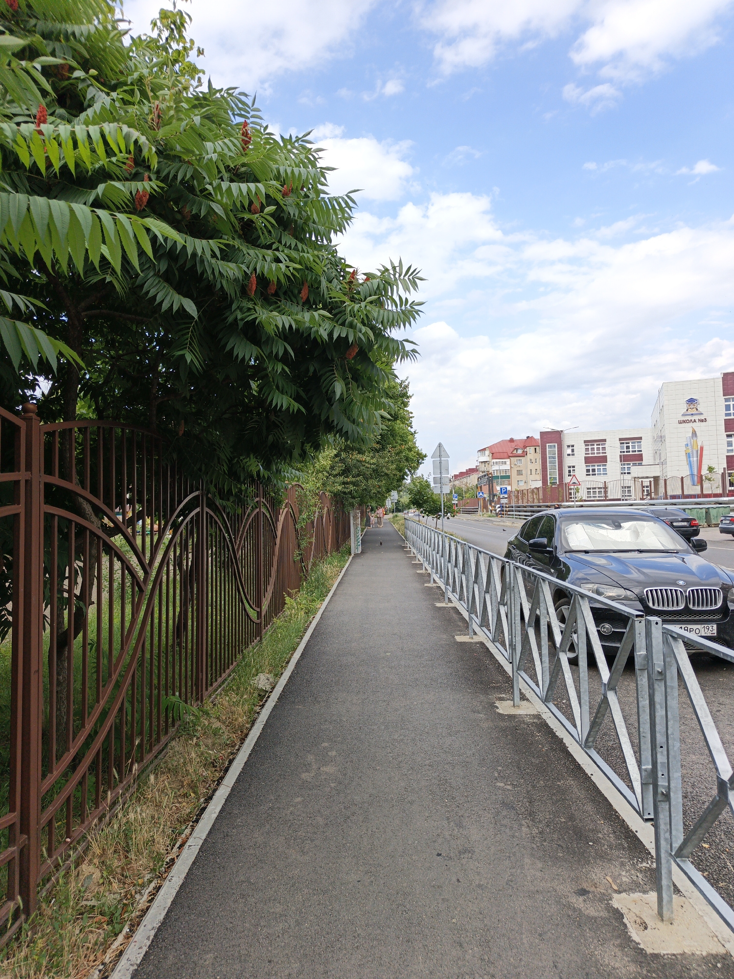 The image depicts a pedestrian sidewalk alongside a street in Goryachiy Klyuch, Russia. On the left, a tall, rusty metal fence with decorative swirling patterns separates the sidewalk from a grassy area and a dense green tree with red, cone-shaped seed clusters. The sidewalk is paved with asphalt and has a concrete curb. On the right side of the sidewalk, a modern, metallic safety barrier runs along the street.
A black SUV is parked next to the sidewalk, facing away from the viewer, with its license plate partially visible. In the background, across the street, there are multi-story buildings, including a school labeled "ШКОЛА №3" (School No. 3). The architecture suggests a residential or institutional area.
The sky is blue with scattered white clouds, indicating fair weather. The lighting suggests it is daytime, likely late morning or afternoon. The scene is quiet, with no prominent activity other than the parked car and a distant figure walking on the sidewalk. The overall mood is peaceful and ordinary, characteristic of a suburban or urban street. The presence of the school and residential buildings gives a sense of community.