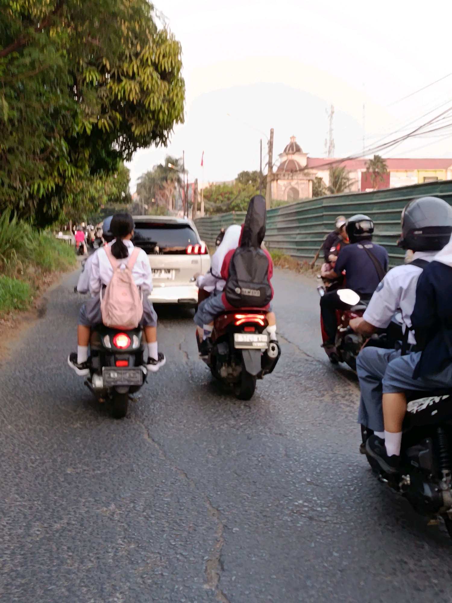 The image depicts a street scene in Bekasi, Indonesia, with several motorcycles and a white car on a paved road. In the foreground, two young people, likely students, are riding on a motorcycle. The person in front, wearing a school uniform and a pink backpack, is seated behind the driver. The driver, also in a school uniform, is operating the motorcycle. Behind them, another motorcycle carries a passenger with a black guitar case strapped to their back. Further down the road, a white car is visible, and other motorcycles and pedestrians are present. The environment suggests an urban or suburban setting with trees lining the left side of the road and buildings and a dome-like structure in the background. The sky is overcast, and the lighting indicates it is either early morning or late afternoon.