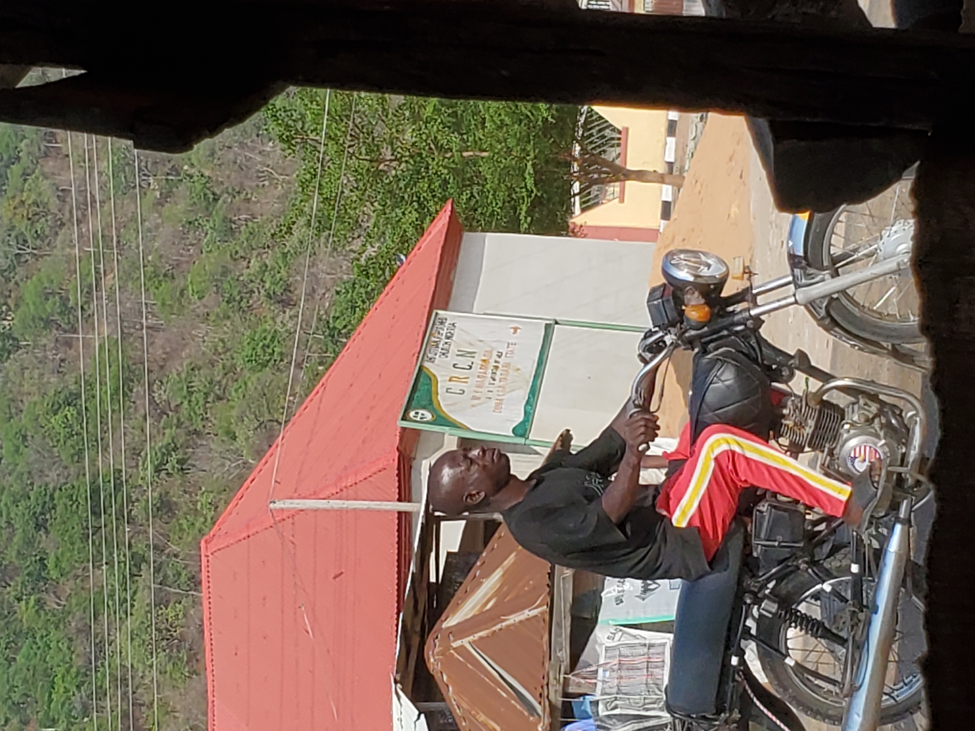 A man with a shaved head is sitting astride a black motorcycle with red and white striped pants, located on a street in Mararaba, Nigeria. He's facing the front of the motorcycle, holding the handlebars, and looking upwards. Behind him is a building with a red roof, and signage for CRCN. The area appears to be outside of a building with a view of trees and other buildings in the background.
