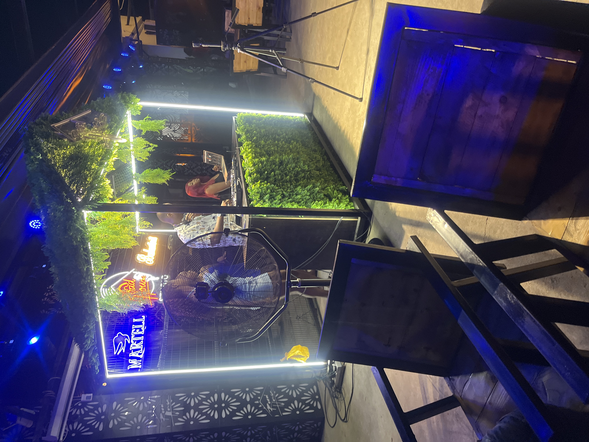The image depicts an outdoor event space, possibly a bar or lounge, in Howayi, Nigeria. In the foreground, two dark wooden chairs with metal frames are positioned facing away from the camera, with a floor that appears to be concrete. The middle ground features a raised platform or booth illuminated by neon lights and adorned with artificial greenery. A large fan is positioned in front of a black structure that may be a DJ booth, with a DJ controller and a laptop visible on the booth. A person with red hair is operating the DJ equipment, and another person in a patterned shirt stands nearby, possibly addressing the crowd. Above the DJ booth, neon signs spell out "MARTELL" and "Road House". In the background, a tripod with a camera is set up, suggesting the event is being recorded. The overall ambiance is that of a vibrant nightlife setting.