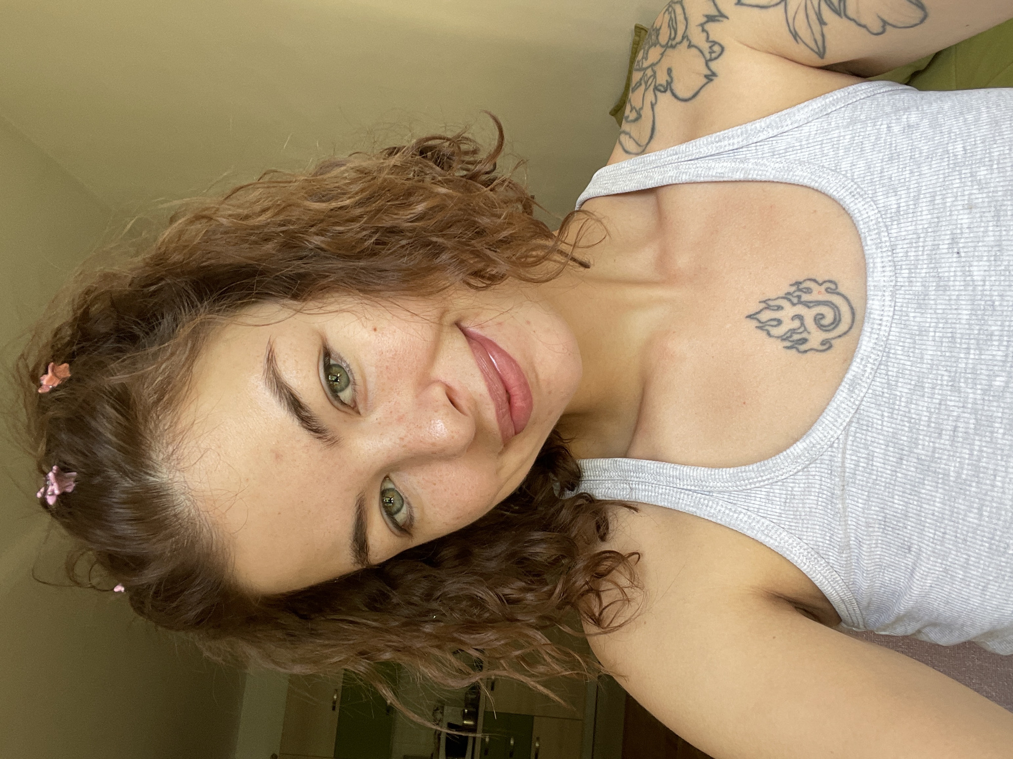 A young woman with curly brown hair, adorned with small pink butterfly clips, poses for a selfie in what appears to be a home environment. Her head is slightly tilted, and she is looking directly at the camera with striking green eyes and a subtle smile. She has a smattering of freckles across her nose and cheeks. She is wearing a grey ribbed tank top, revealing a tattoo of flames on her chest and floral designs on her left shoulder and arm. The background is softly lit, with a hint of a kitchen with cabinets visible on the left side and a plain wall on the right. The overall impression is casual and relaxed. The image does not provide any information to suggest it is located in Boreti, Montenegro, or any specific city.