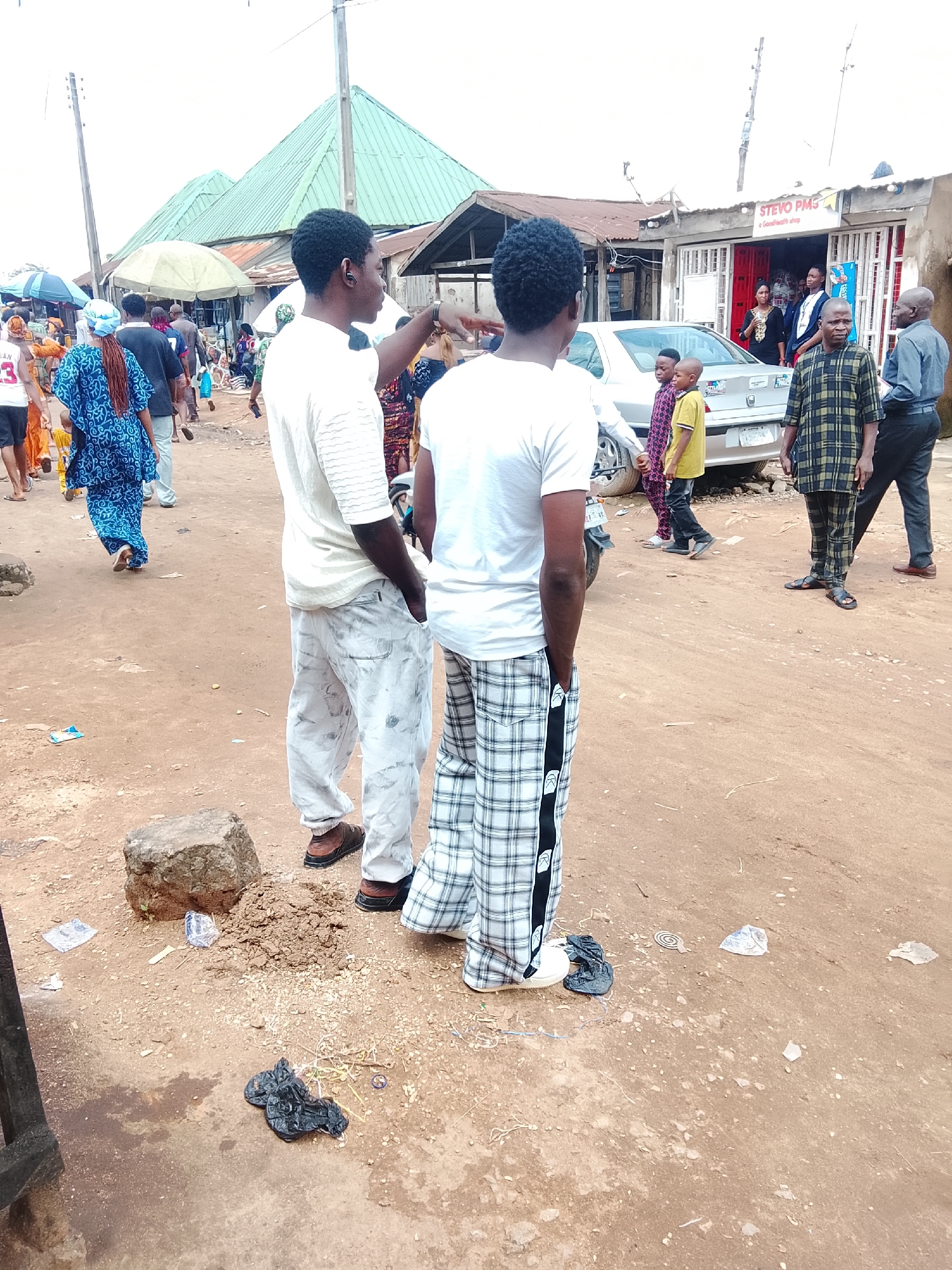 This image depicts a street scene in Abuja, Nigeria, likely a market or commercial area. Two young men, one in a white t-shirt and light-colored, patterned pants, and the other in a white t-shirt and light gray pants, are the central figures. The man on the left is gesturing with his right hand. Several other individuals are visible in the background, including children, women in traditional attire, and men in various casual and formal wear. A silver car is parked on the right side of the image. The environment is dusty with a dirt road and scattered debris. Several market stalls with green roofing are visible in the background, suggesting a commercial hub. The sky is overcast.