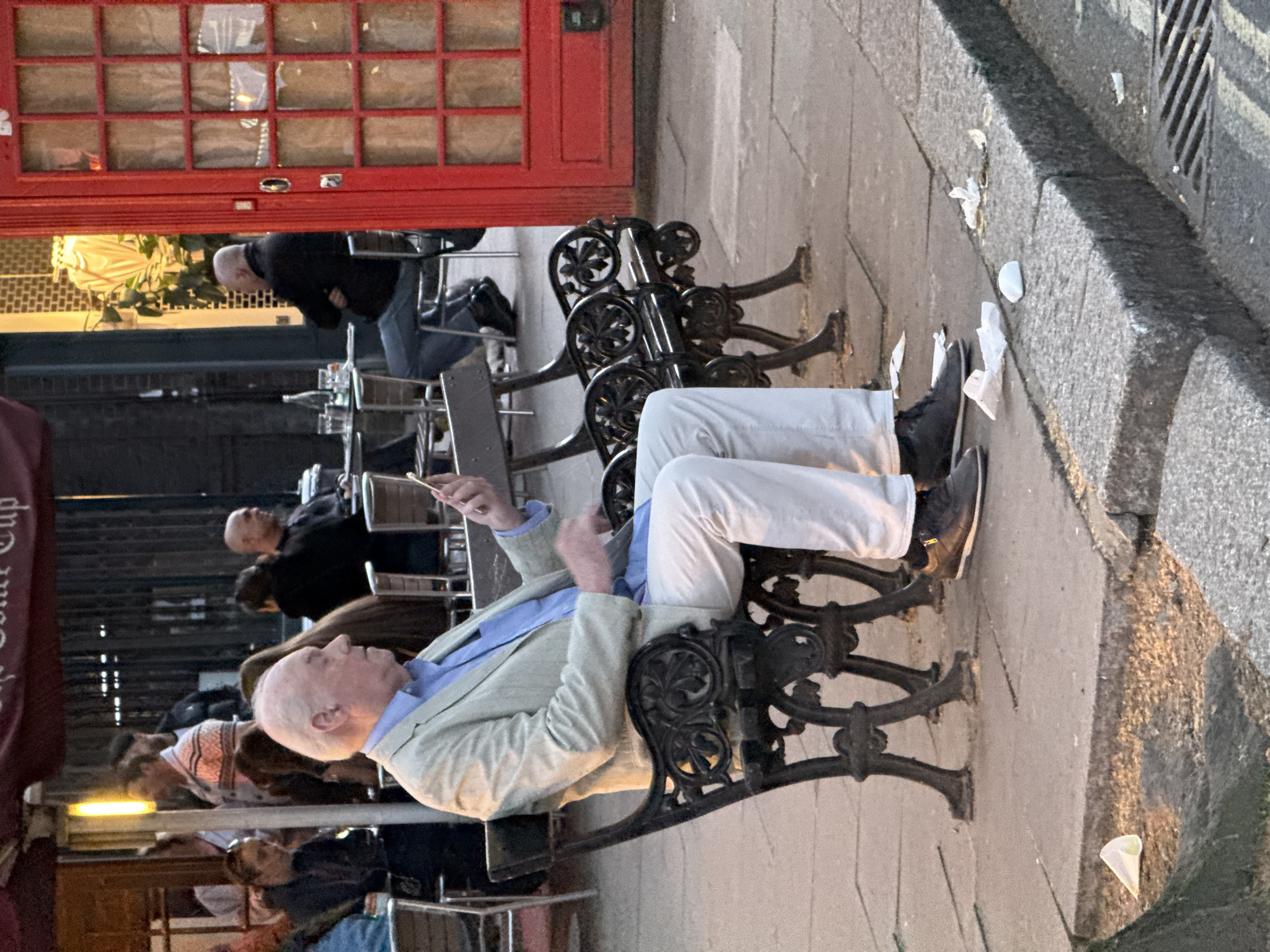 An eye-level, medium shot depicts a man sitting on a black, wrought-iron bench along a sidewalk. He wears a light green jacket, a blue shirt, and white pants. His shoes are dark gray. He holds a small, brown object in his hand and appears to be looking at it. Other people are visible in the background, seated at tables outside what appears to be a business. A red door or window frame is visible on the left, and part of a striped awning is seen above the tables. Litter is scattered on the sidewalk beside the bench. The photo appears to have been taken in London, United Kingdom.