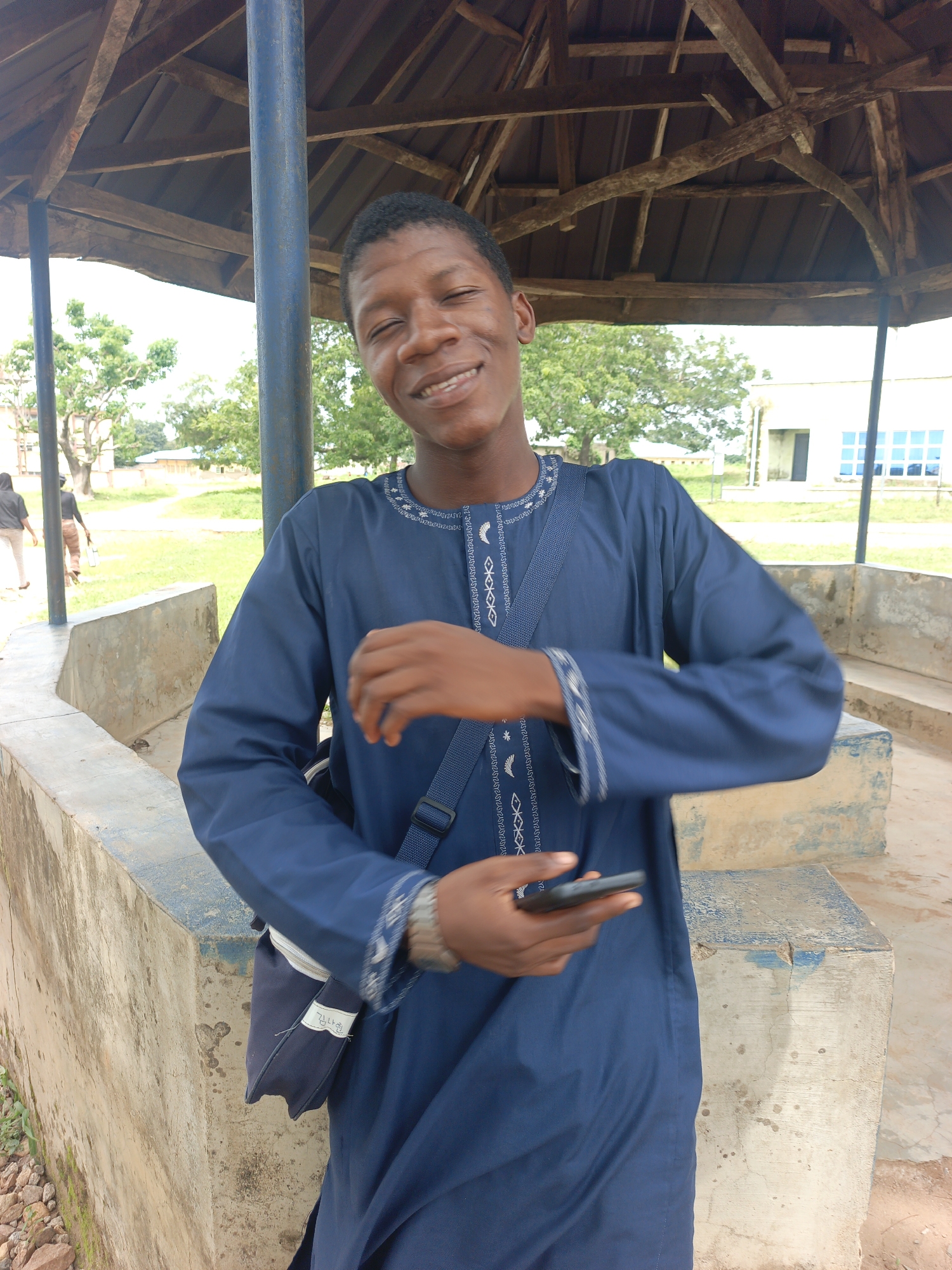 A young Black man wearing a dark blue tunic with white embroidery is standing under a wooden shelter. He is smiling and holding a smartphone in his right hand, which is raised. A dark blue messenger bag with white stitching and a small white tag with Korean characters is slung across his body. He is positioned in front of a concrete structure with blue accents. In the background, there is a grassy area with trees and a building with blue windows. Two other people, one in black clothing and another in striped pants, are walking in the distance. The overall setting appears to be outdoors in a public space, possibly a school or community area, located in Akwanga, Nigeria.