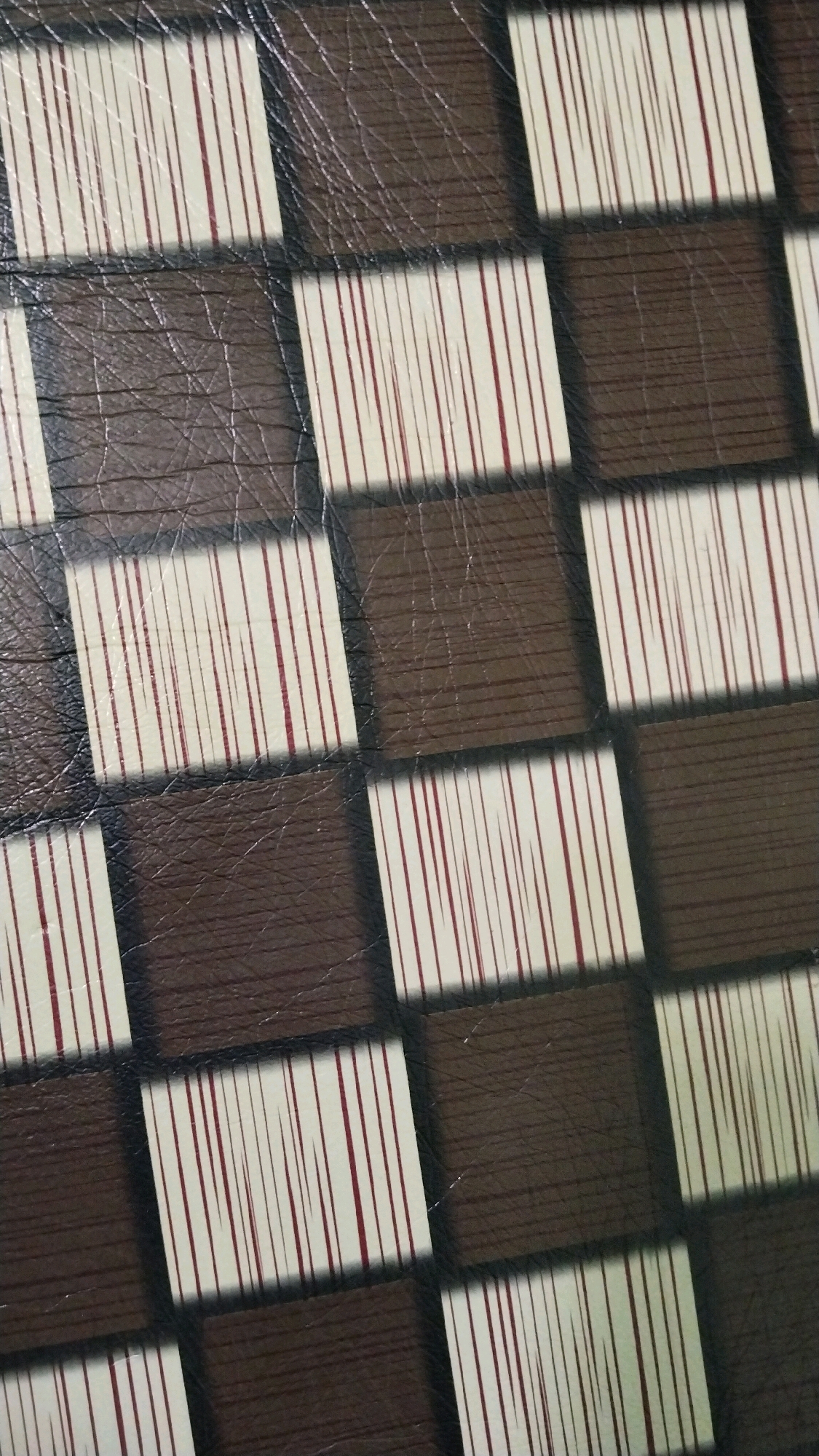 This is a close-up, overhead shot of a textured surface resembling a checkerboard pattern. The squares alternate between dark brown, with a faux leather or wood grain texture and visible creases, and off-white with thin vertical red lines. The lines within the off-white squares are irregular and vary in thickness. The lighting casts subtle shadows that highlight the texture and the edges of the squares. The image does not contain any discernible elements that would place it in a specific location in Simo, Indonesia.