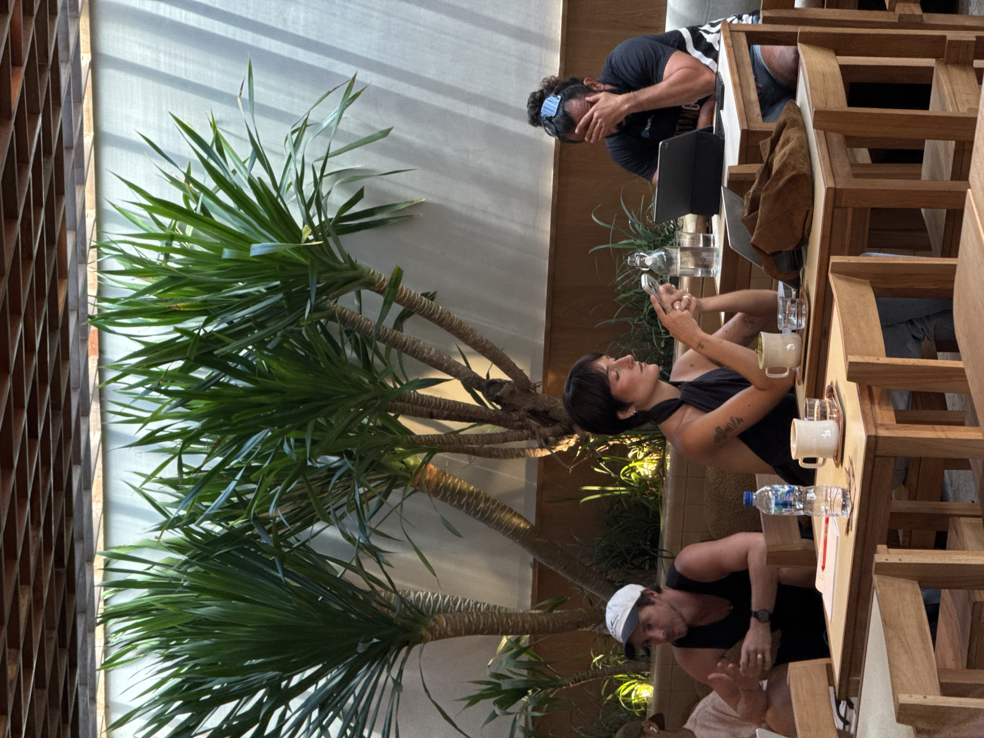 The image depicts three individuals at an outdoor cafe in what appears to be North Kuta, Indonesia. The environment includes decorative palm trees next to a building with a trellis. A man is seated at the end of the table, looking down at a tablet. A woman sits in the middle and is holding her cell phone, while the man on the other end is looking toward her and gesturing with his hand. There are drinks and electronic devices on the wooden table.