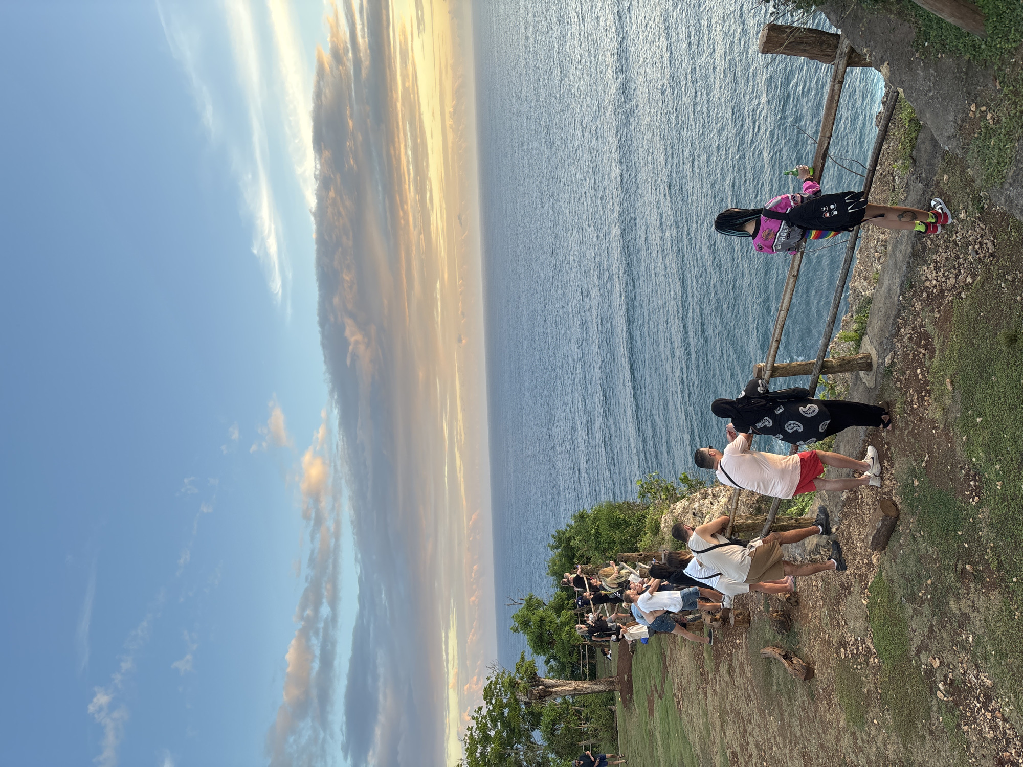 Here's a description of the image:
The image captures a scene at Uluwatu Temple in Kuta Selatan, Indonesia. Numerous people are lined along the cliff edge, seemingly observing a sunset over the ocean. A wooden railing borders the cliff. The sky features a gradient of blues and sunset colors. The ocean stretches to the horizon.