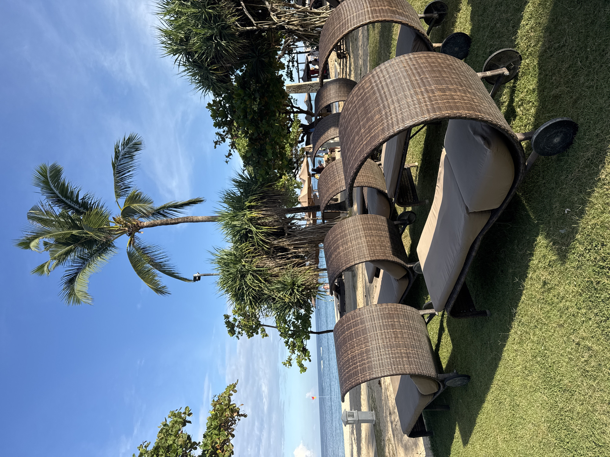 The image shows a beachside scene in Kuta Selatan, Indonesia. A row of lounge chairs with woven canopies are lined up on a grassy area next to a sandy beach. A palm tree and other tropical vegetation are visible in the background. The sky is blue with scattered clouds. In the distance, the ocean can be seen with a small red flag marking its edge.