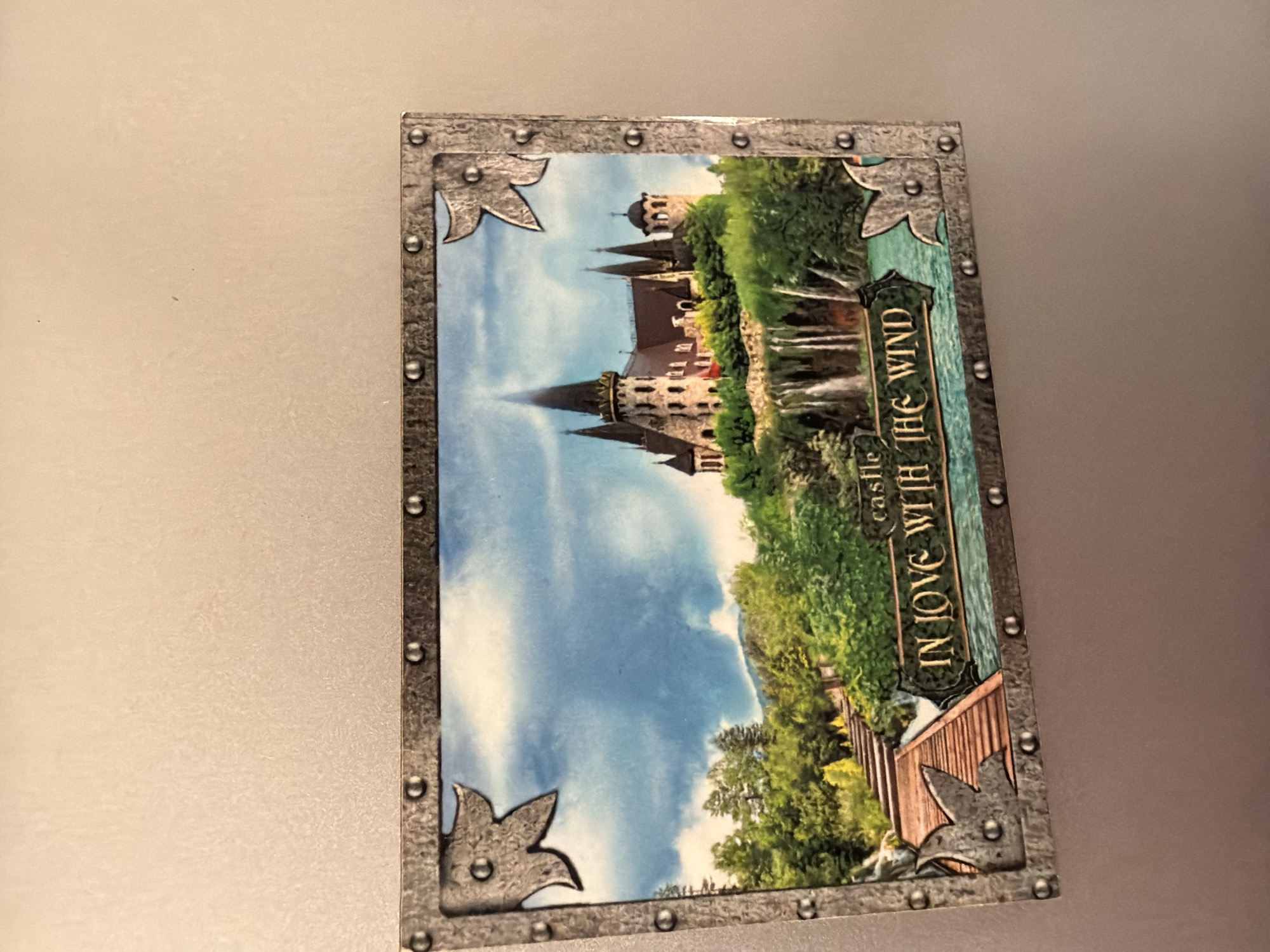 The image is a postcard depicting a castle scene. The postcard has a metallic border with decorative corner pieces and studs. The castle is a large, ornate structure with multiple spires and towers, perched on a cliff overlooking a waterfall and a body of water. Lush green trees and foliage surround the castle, and the sky is cloudy blue. Text on the postcard reads "castle IN LOVE WITH THE WIND." The postcard is not located in the city of Pazardzhik, Bulgaria.