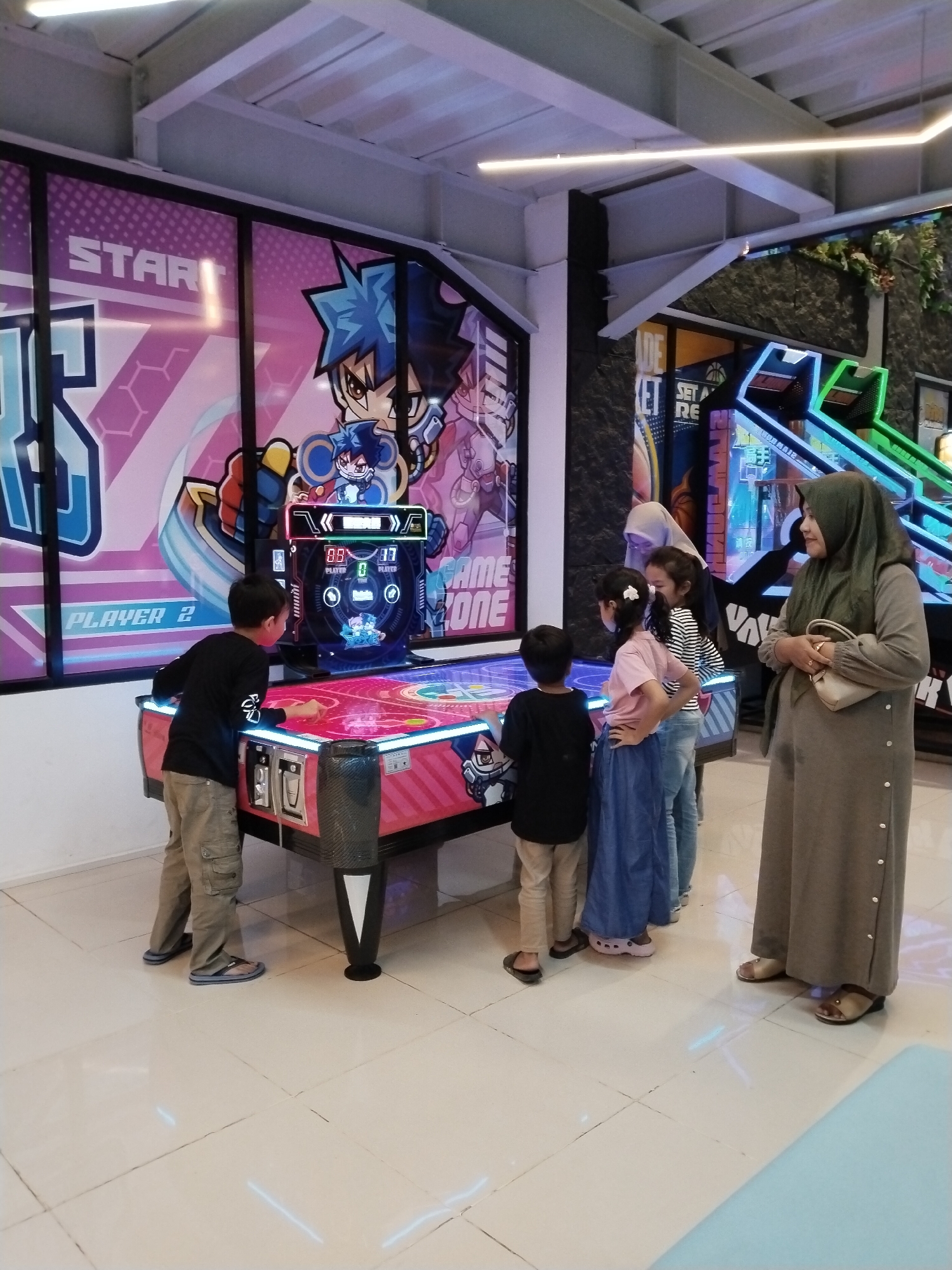 In a brightly lit indoor arcade located in Kecamatan Pati, Indonesia, a group of children and adults are gathered around an air hockey table. The air hockey table itself is a prominent pink and blue object with neon lights, displaying scores of "88" and "17" for "Player 1" and "Player 2" respectively. The backdrop of the arcade features large graphics with cartoon characters and the words "START" and "GAME ZONE."
A young boy in a black long-sleeved shirt and khaki pants stands on the left, facing the air hockey table and appears to be playing. To his right, several other children are also observing or participating. One girl in a pink shirt and blue skirt stands with her hands on her hips, and another in a striped shirt and jeans is next to her. Behind them, a woman wearing a light grey hijab and a long grey dress, holding a beige handbag, stands and watches. Another woman, partially visible in a grey hijab, is also present. The floor is tiled with a light-colored, reflective surface. In the background, other arcade machines with neon lights are visible, suggesting a lively entertainment venue.