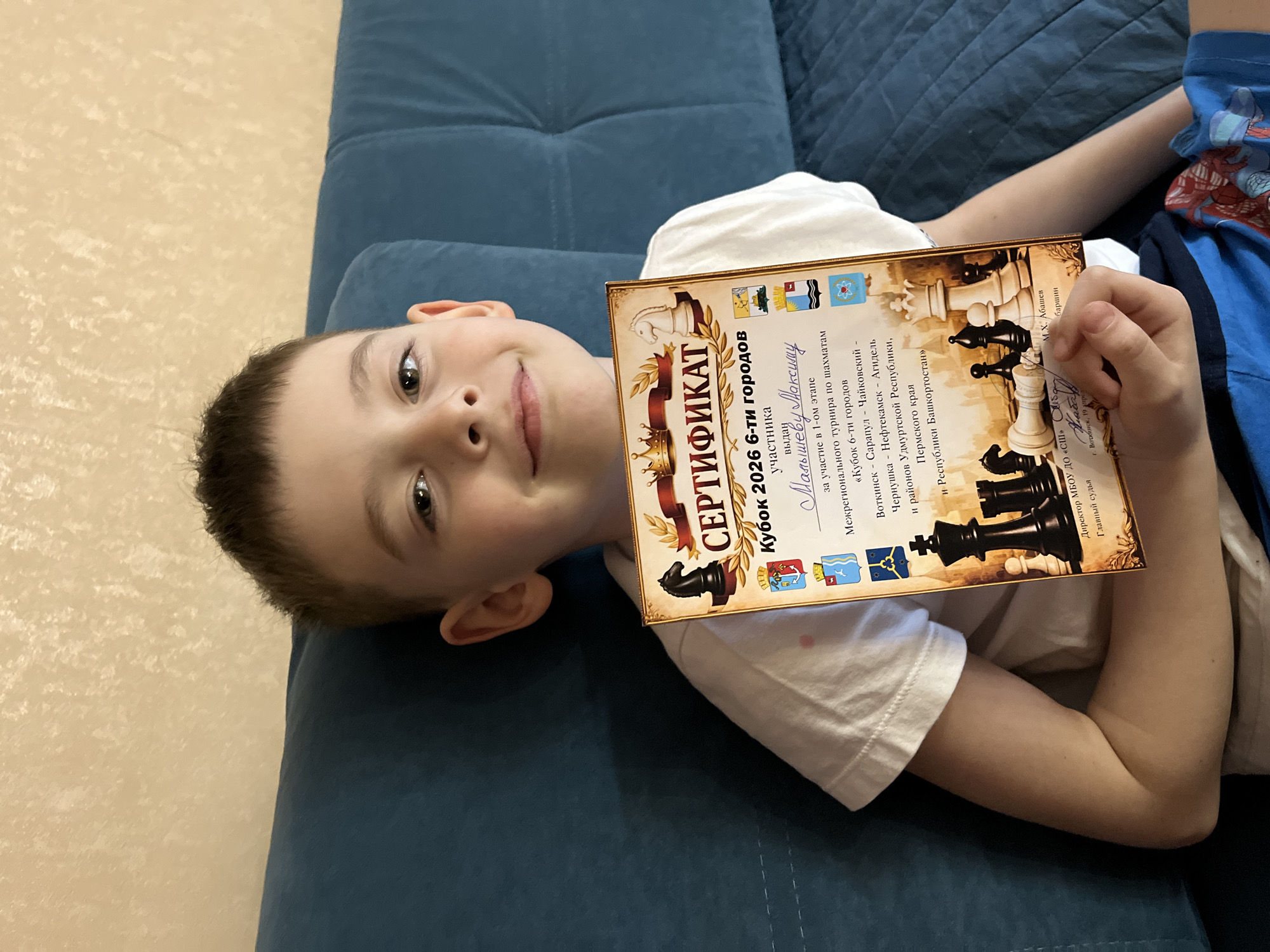 A young boy is lying on a blue couch while holding up a certificate. The certificate states that it is for the "Cup of 2026 6 cities" chess tournament. The boy's name is listed as "Malyshev Maxim." The tournament took place in the cities of Votkinsk, Sarapul, Chaikovsky, Chernoushka, Neftekamsk, and Agidel, as well as the regions of the Udmurt Republic, the Perm region, and the Republic of Bashkortostan. The Director is M.Kh. Abashev. The city of Votkinsk is listed on the certificate. The name of the city of Chaikovsky, Russia, appears on the certificate.