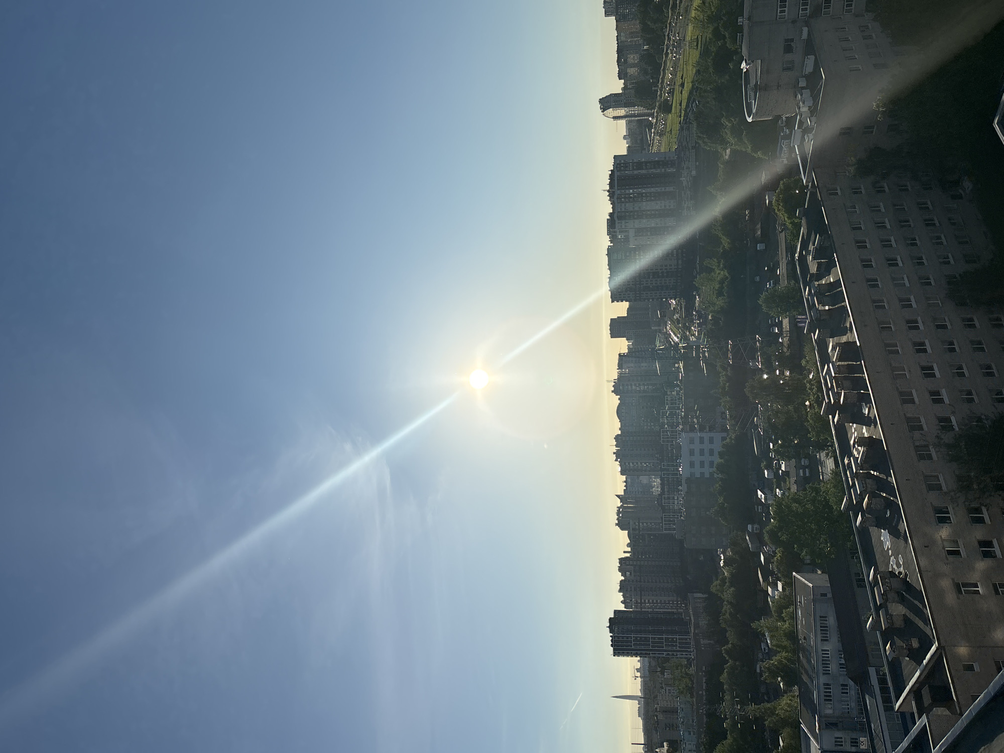 The image depicts a cityscape of Saint Petersburg, Russia, under a clear blue sky with a prominent sun flare diagonally crossing the frame from the upper left to the lower right. The sun is bright and positioned in the upper middle of the sky, creating a lens flare effect. In the distance, a cluster of modern high-rise buildings forms the skyline, with a particularly tall, slender skyscraper visible on the far left. Closer to the foreground, several mid-rise apartment buildings with numerous windows are visible. Green trees dot the landscape between the buildings, and a network of roads and what appears to be a parking lot with several vehicles are present. The foreground shows the roofs of some older apartment buildings with multiple chimneys. The overall scene suggests an urban environment on a sunny day.
