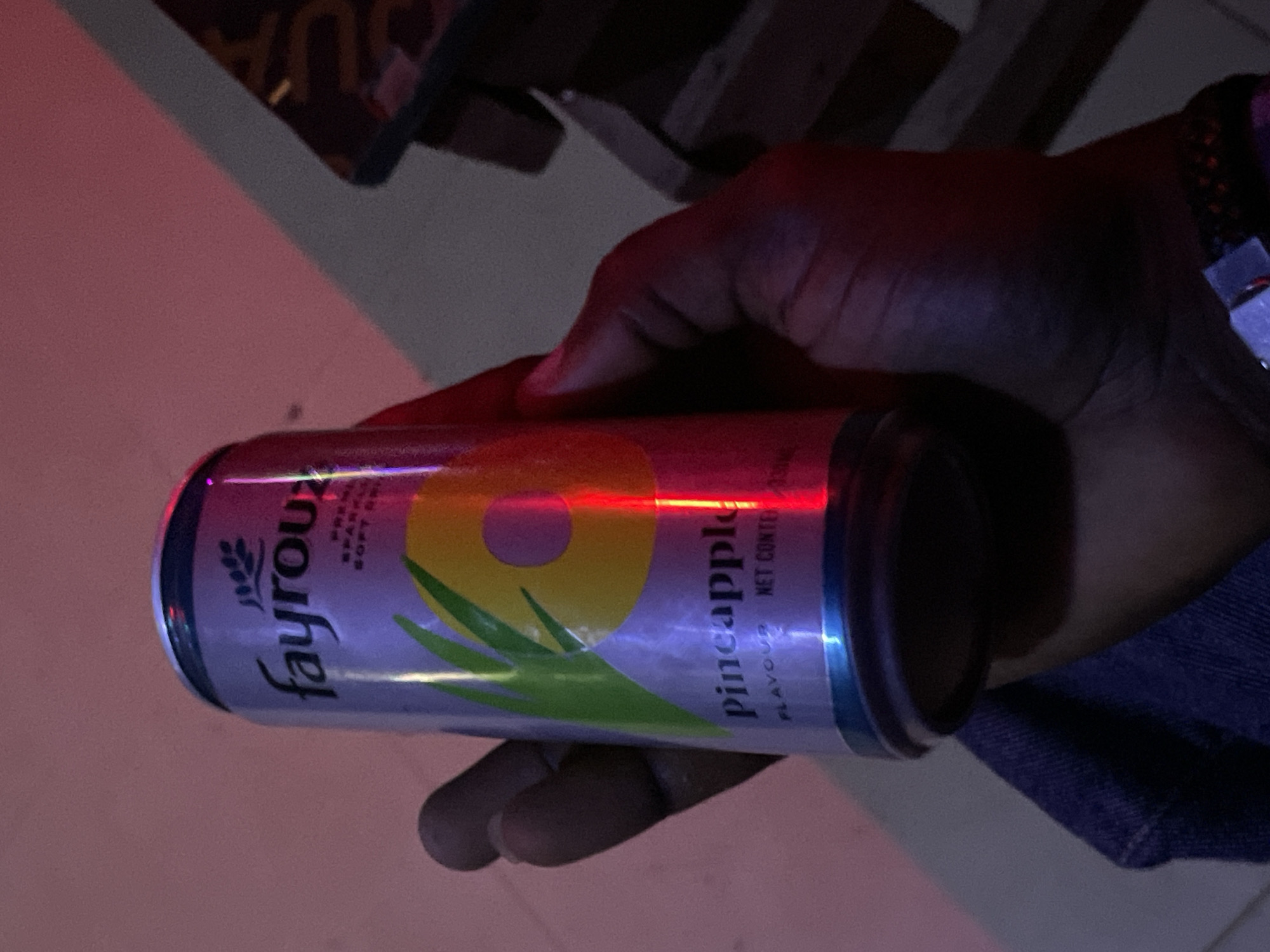 The image shows a person's hand holding a can of Fayrouz pineapple-flavored soft drink. The can is primarily silver with a design featuring a pineapple and green leaves. The word "pineapple" is visible in large text on the can. The lighting is dim with red and purple hues, suggesting an indoor environment, possibly a bar or lounge. A portion of the person's dark-skinned hand and arm is visible, along with a dark blue denim jacket. The background is out of focus and shows dimly lit furniture, consistent with a social setting. There is no indication of the specific location in Durimi, Nigeria, beyond it being a place where such a drink would be consumed.