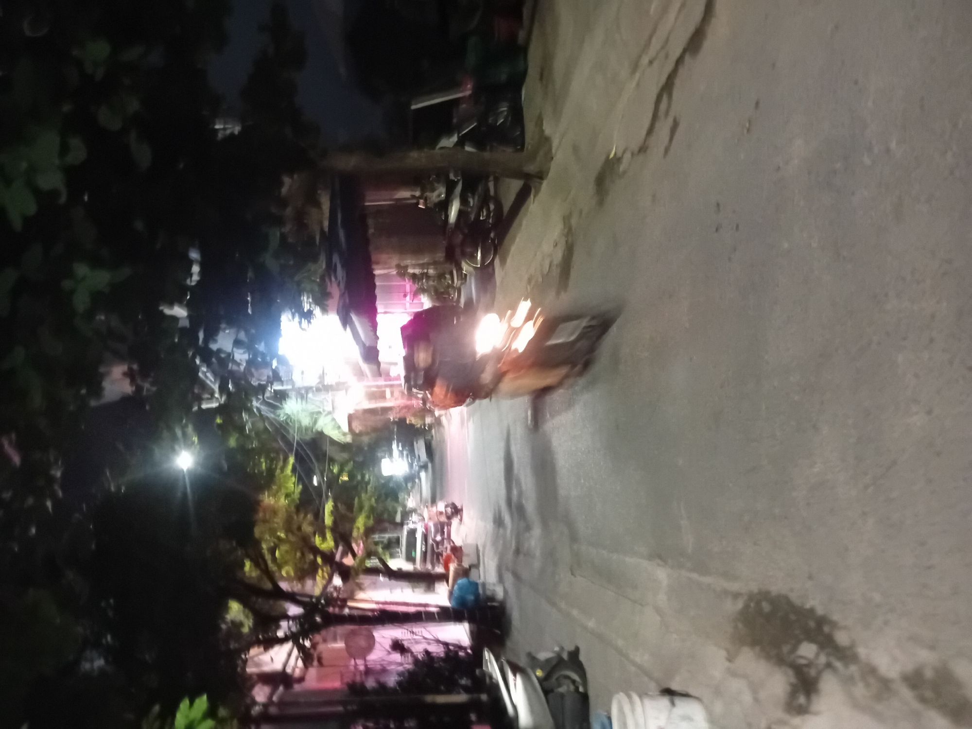 The image depicts a street scene at night in Da Nang, Vietnam, likely a residential or commercial alley. A person on a scooter, with their headlight illuminated, is captured in motion, their form blurred by movement. The street is paved and appears wet in places, reflecting the ambient light from streetlamps and the scooter's headlight. Trees with dark foliage overhang the street, casting shadows. Buildings line the sides of the street, some with illuminated windows and awnings. Parked scooters and what appear to be buckets or containers are visible along the side of the street. The overall atmosphere is dimly lit and captures a typical evening streetscape.