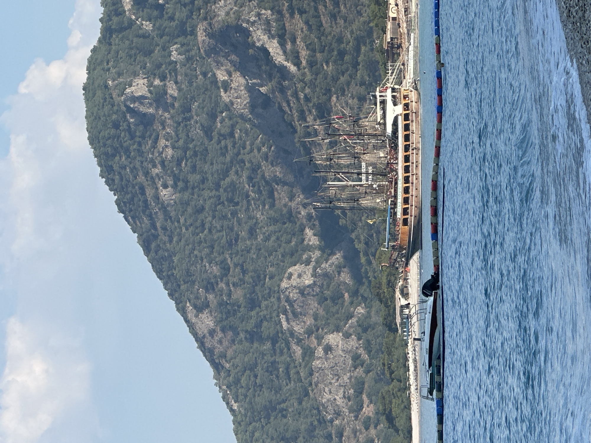 A wide shot captures a scenic coastal view with a lush, tree-covered mountain dominating the background. The mountain is rocky in places and appears to be a significant natural feature. In the middle ground, several boats are anchored in the clear blue water. A large, multi-masted sailing ship, possibly a pirate ship replica, is prominently positioned, with its sails furled. Smaller boats, including a white speed boat, are also visible. A floating barrier of colorful buoys separates the boats from the foreground water. The foreground shows the calm, blue sea with gentle waves lapping at a pebbled shore. The sky is bright blue with scattered white clouds, indicating a sunny day. The overall scene suggests a popular tourist destination, likely Fethiye, Turkey, given the characteristic landscape and the presence of leisure boats.