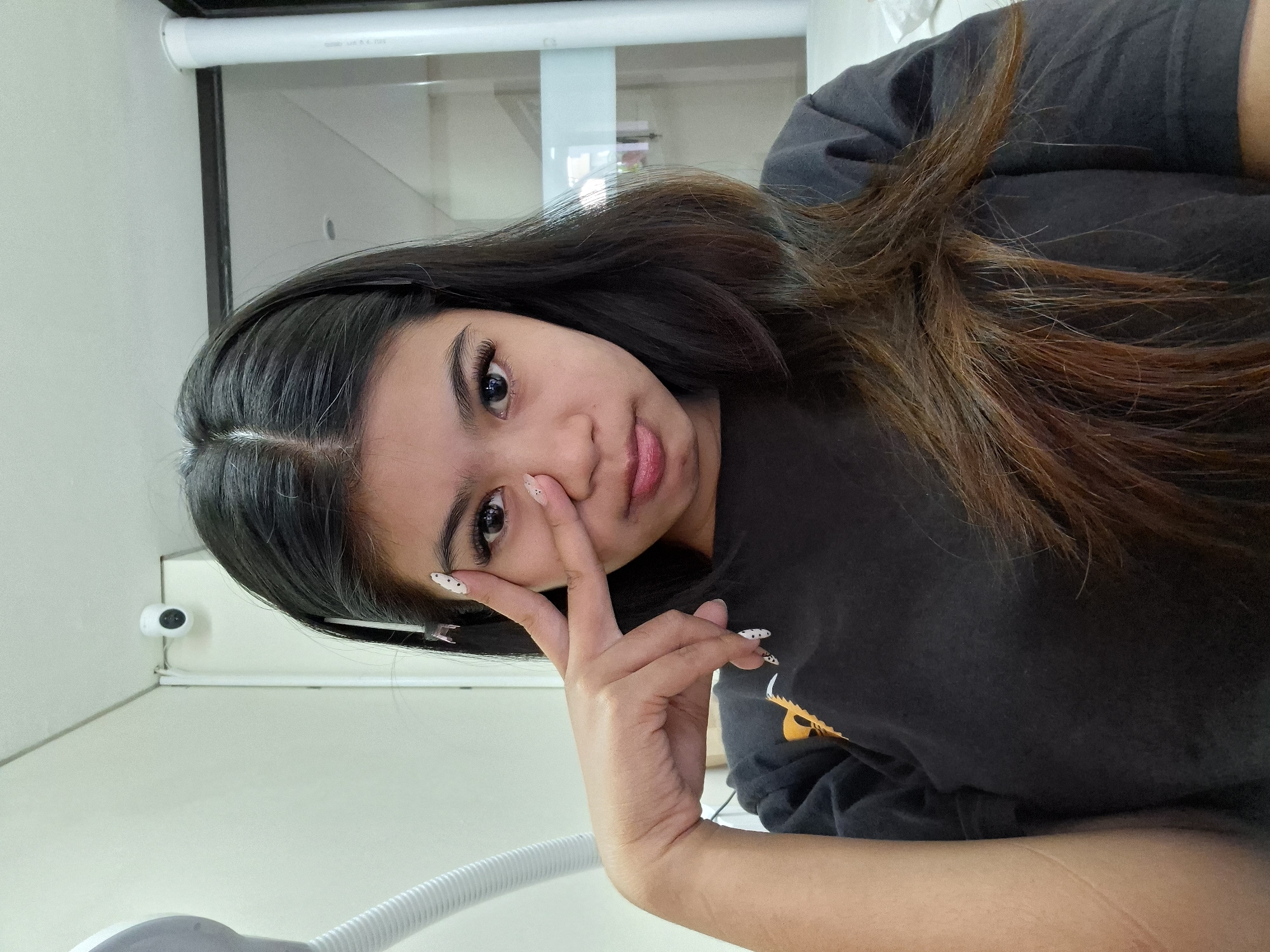 A young woman with long, dark brown hair is pictured in a close-up, self-taken photo. She is wearing a dark grey t-shirt with a yellow graphic on the left side of her chest. Her right hand is raised, with her index and middle fingers extended in a peace sign, partially covering her right eye. She is looking directly at the camera with a neutral expression, her lips slightly parted. Her fingernails are long and painted white with black polka dots. In the background, a white ceiling is visible, with a small white security camera mounted in the upper left corner. To the right, a portion of a white vertical pipe and a glass partition are discernible. The overall lighting suggests an indoor setting. The image is tagged with Kuta, Indonesia, indicating the possible location.