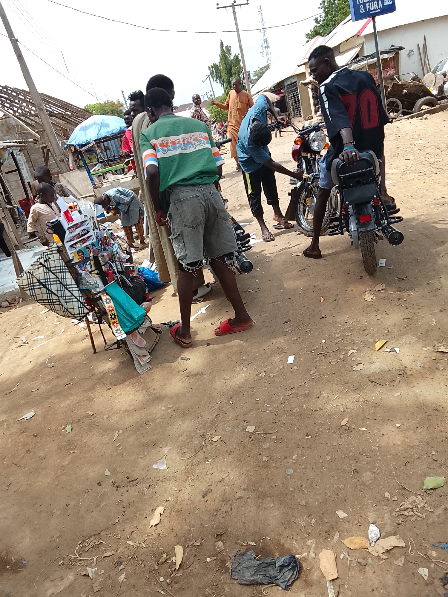 The image shows a street scene in Abuja, Nigeria. Several people are gathered around a motorcycle and a small vendor stall. One man in a blue hoodie is inspecting the engine of the motorcycle, while another man in a black and white shirt with the number 70 is sitting on the motorcycle. A man in a green shirt with the word "GUESS" on it stands nearby. A vendor stall displays various items, including sunglasses, bags, and trinkets. The ground is dirt with scattered debris. The sky is overcast. A sign for "Yogurt & Fura" is visible in the background.