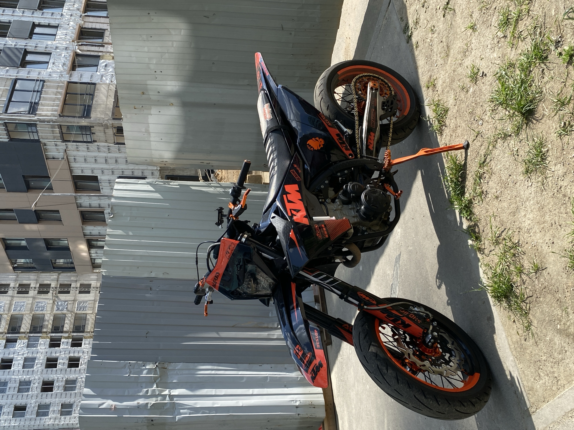 The primary subject is an orange and black KTM motorcycle, appearing to be a supermoto or off-road style bike. No people are visible.
The motorcycle is parked outdoors in what appears to be an urban or construction environment. On the left side of the image, a tall building under construction or renovation is visible, featuring multiple windows and insulation material on its exterior. A corrugated metal fence or hoarding stands between the building and the motorcycle. On the right side, there's a concrete path with patches of sparse green grass and dirt.
The motorcycle is stationary, parked on its orange kickstand. There is no active event taking place. The time of day is bright daylight, indicated by strong shadows cast by the motorcycle. The weather appears clear and sunny.
The motorcycle features prominent orange detailing, including its wheels, frame, kickstand, and handlebar levers. Several parts, such as the exhaust and some body panels, have a black carbon fiber texture. The ground around the bike is dusty.
Visible text and logos include: "KTM" prominently on the fuel tank, side panels, and wheels; "ACERBIS" on the front fender; "BRAKING" on the rear swingarm; "DUNLOP" on the front tire; a partial "AKRAPOVIC" on the exhaust; a stylized lion/animal head logo on a side panel; and "SMC 690" on the front fender. The building's insulation material shows repeating text "I_TEN".