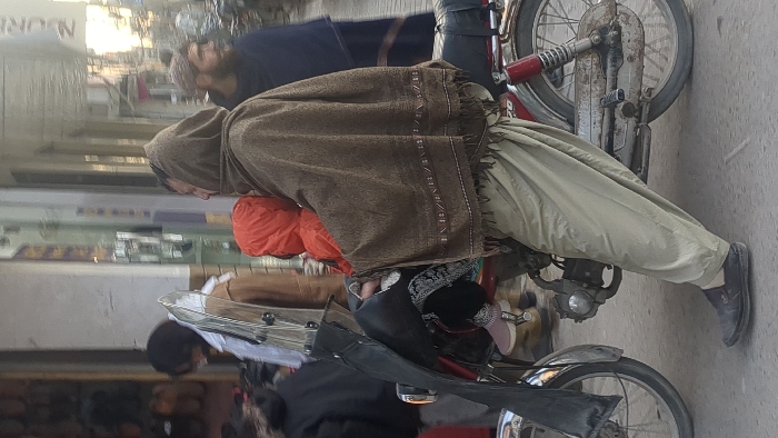 The image depicts a street scene in Quetta City Tehsil, Pakistan, under daylight conditions. In the foreground, a male individual is seated on a red Honda CD 70 motorcycle, balancing with his right foot on the pavement. He wears a dark brown shawl draped over his head and shoulders, light olive-green shalwar, and dark slip-on shoes. Seated in front of him on the motorcycle's fuel tank is a small child, wearing an orange hooded jacket, dark pants with white embroidered patterns, and light pink/beige shoes with white soles. The motorcycle has a clear windshield and a black leather handlebar cover. To the left, a retail storefront displays various footwear. A person with dark hair, wearing a white shirt over a light brown jacket, stands near the store entrance, partially obscured. Another individual, seen from the back in a dark jacket, is positioned to the left of the motorcycle's front wheel. To the right, another male individual with a beard and a light-colored cap wears dark blue clothing and faces away from the camera. The background shows blurred outlines of urban buildings and street activity.