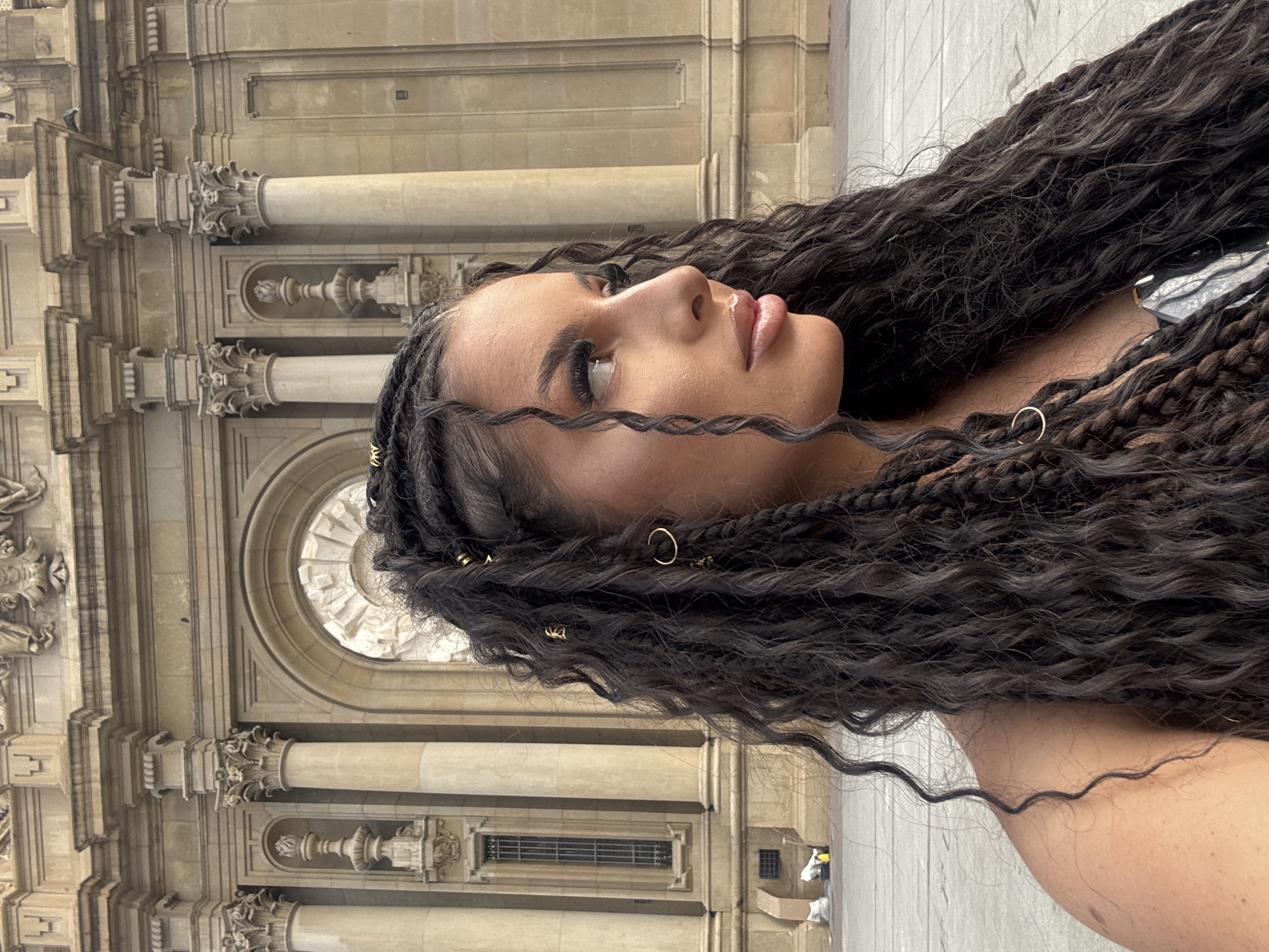 In Zaragoza, Spain, a young woman with braided dark hair adorned with gold accessories stands in front of the Basilica of Our Lady of the Pillar. The woman is positioned in the foreground, facing right in profile. Her makeup is visible, with emphasis on her eyes and lips. Behind her, the grand facade of the basilica, characterized by its Baroque architecture, ornate columns, and arched windows, dominates the background. The plaza in front of the basilica is paved and appears to be a public space. The lighting suggests it is daytime.