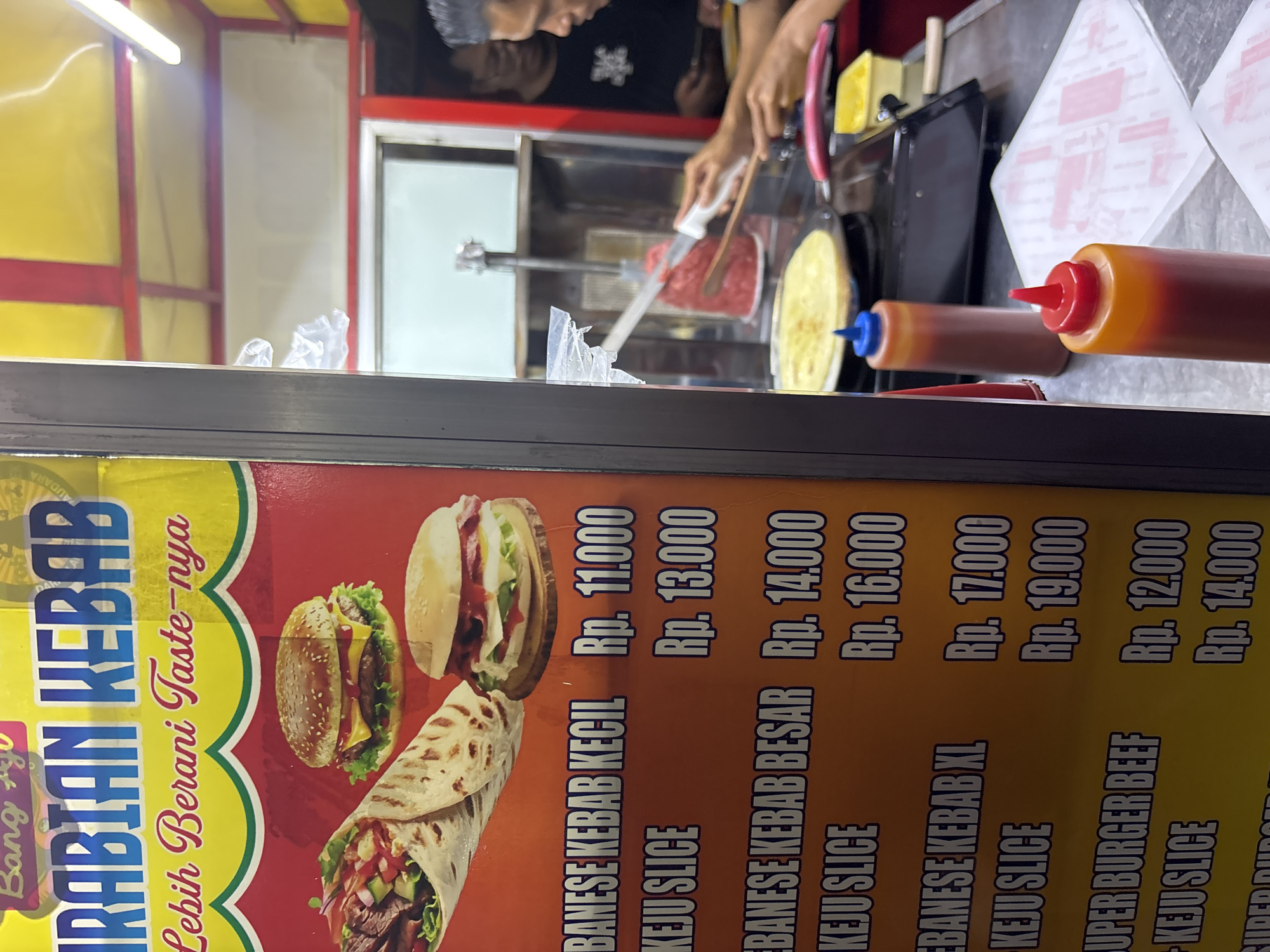 The image shows a street food stall in North Jakarta, Indonesia, specializing in kebabs and burgers. A large, colorful sign for "ARABIAN KEBAB" with the slogan "Lebih Berani Taste-nya" (Bolder Taste) is prominently displayed. The sign features images of a kebab wrap and burgers. Below the images, a menu lists prices in Indonesian Rupiah (Rp.) for various items like "BANESE KEBAB KECIL" (Small Banese Kebab) for Rp. 11,000, "KEJU SLICE" (Cheese Slice) for Rp. 13,000, "BANESE KEBAB BESAR" (Large Banese Kebab) for Rp. 14,000, and "KEJU SLICE" for Rp. 16,000. Further down, "SUPER BURGER BEEF" is priced at Rp. 12,000 with "KEJU SLICE" for Rp. 14,000.
In the background, a person is preparing food, likely a crepe or flatbread, on a griddle. Another person, with dark hair and wearing glasses, is visible in the stall. Two squeeze bottles, one with a red cap and the other with a blue cap, are on the counter, suggesting they contain sauces. The overall environment appears to be an open-air food stall with basic equipment.