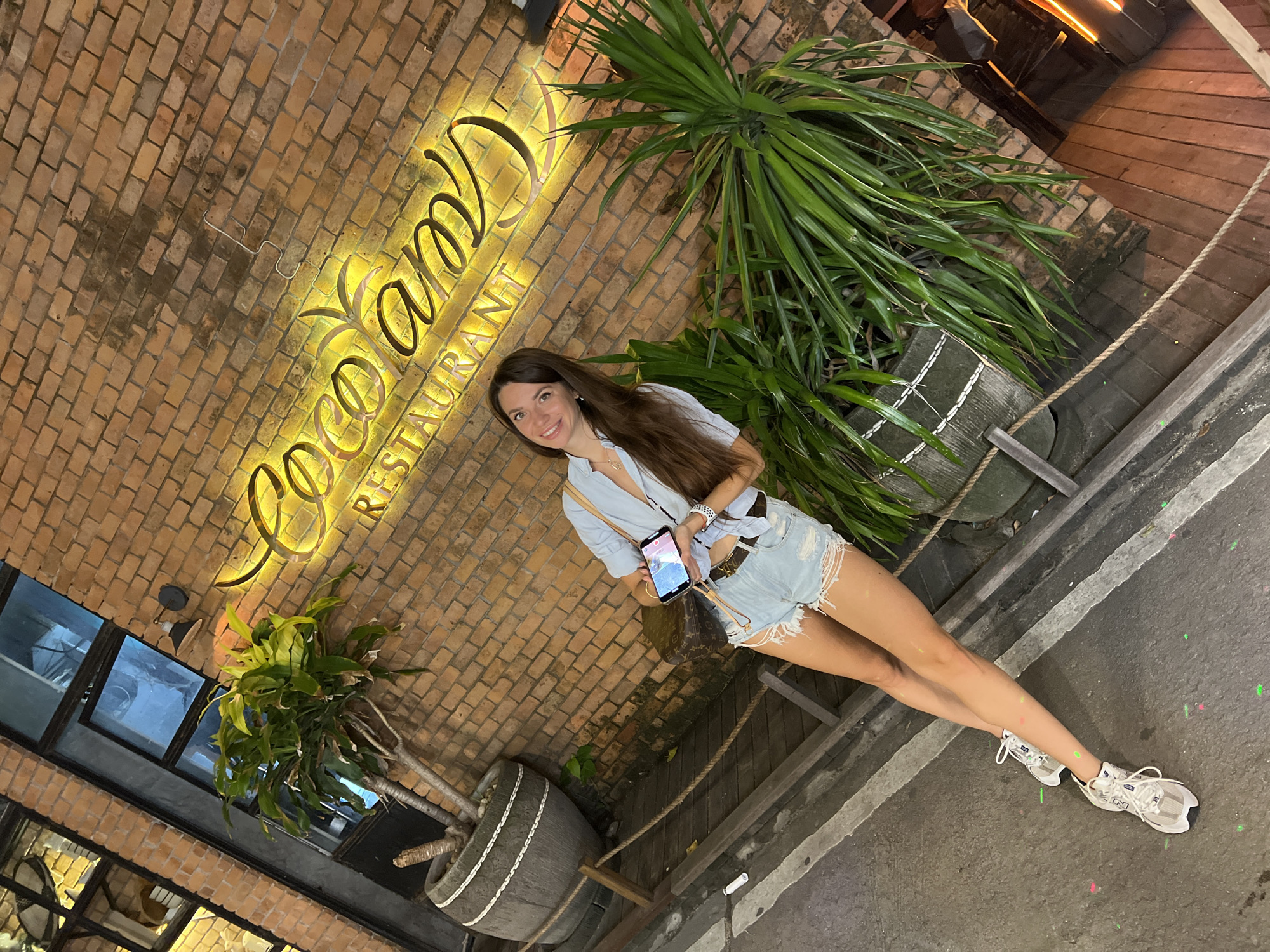 A full shot, taken at night, shows a smiling, long-haired woman, posing near the illuminated "Coco Tam's Restaurant" sign in Baan Bo Phut, Thailand. She is wearing a light blue top and denim shorts, holding a cell phone in her left hand. She is positioned at a slight angle. Behind her is a brick wall, the restaurant sign, and two planters with green plants. The ground is a mix of stone and concrete.
