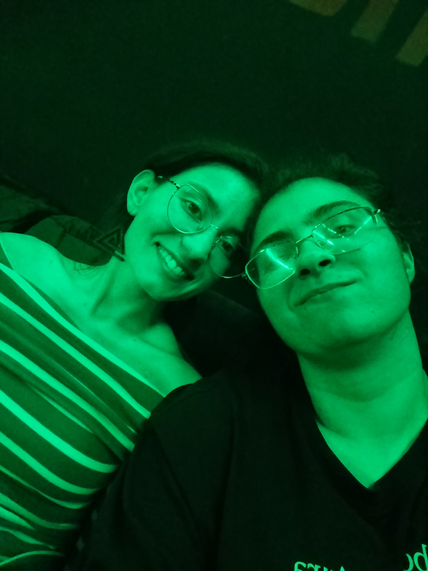 Two individuals are captured in a selfie-style photograph under green lighting. The person on the left wears glasses and a horizontally striped top, while the person on the right also wears glasses and a dark v-neck shirt with text partially visible at the bottom. The setting is indoors, and the image is likely taken in Sofia, Bulgaria.