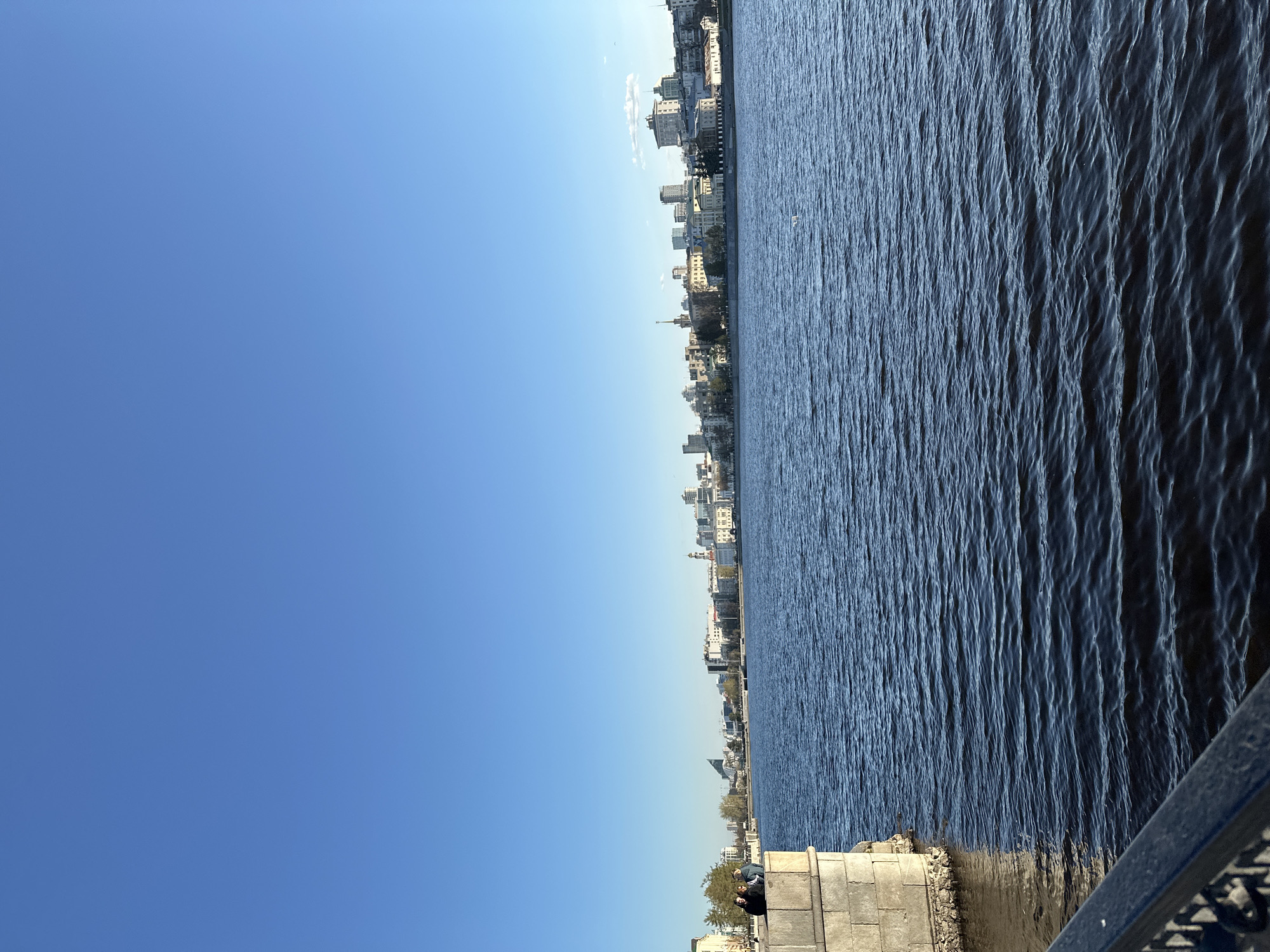 The image, correctly oriented, captures a panoramic view of an urban waterfront scene under a bright, clear blue sky. The upper half of the frame is dominated by an expansive, cloudless sky, displaying a gradient of blue hues.
Along the mid-line of the image, stretching horizontally, is the distinct skyline of a city. It comprises a mix of buildings, including multi-story structures with varied architectural styles, some featuring intricate details or spire-like elements, suggesting both historical and modern construction. These buildings form a continuous band between the sky and the water.
The lower half of the image is filled by a wide body of dark blue water, likely a river or a large lake, exhibiting numerous small ripples and waves across its surface, indicating movement. The water's texture is detailed, reflecting ambient light.
In the bottom right corner, a robust stone embankment or quay is visible, with a dark metal railing partially entering the frame. Two individuals are seated on this stone structure, partially obscured by some sparse vegetation, appearing to be observing the water or the distant cityscape.
The scene portrays a sunny day, likely late morning or early afternoon, given the even and bright lighting, with no visible clouds or strong, elongated shadows. The weather appears calm and clear. The setting depicts a harmonious blend of urban infrastructure and natural water features, creating a serene atmosphere in Yekaterinburg, Russia. There is no visible text in the image.