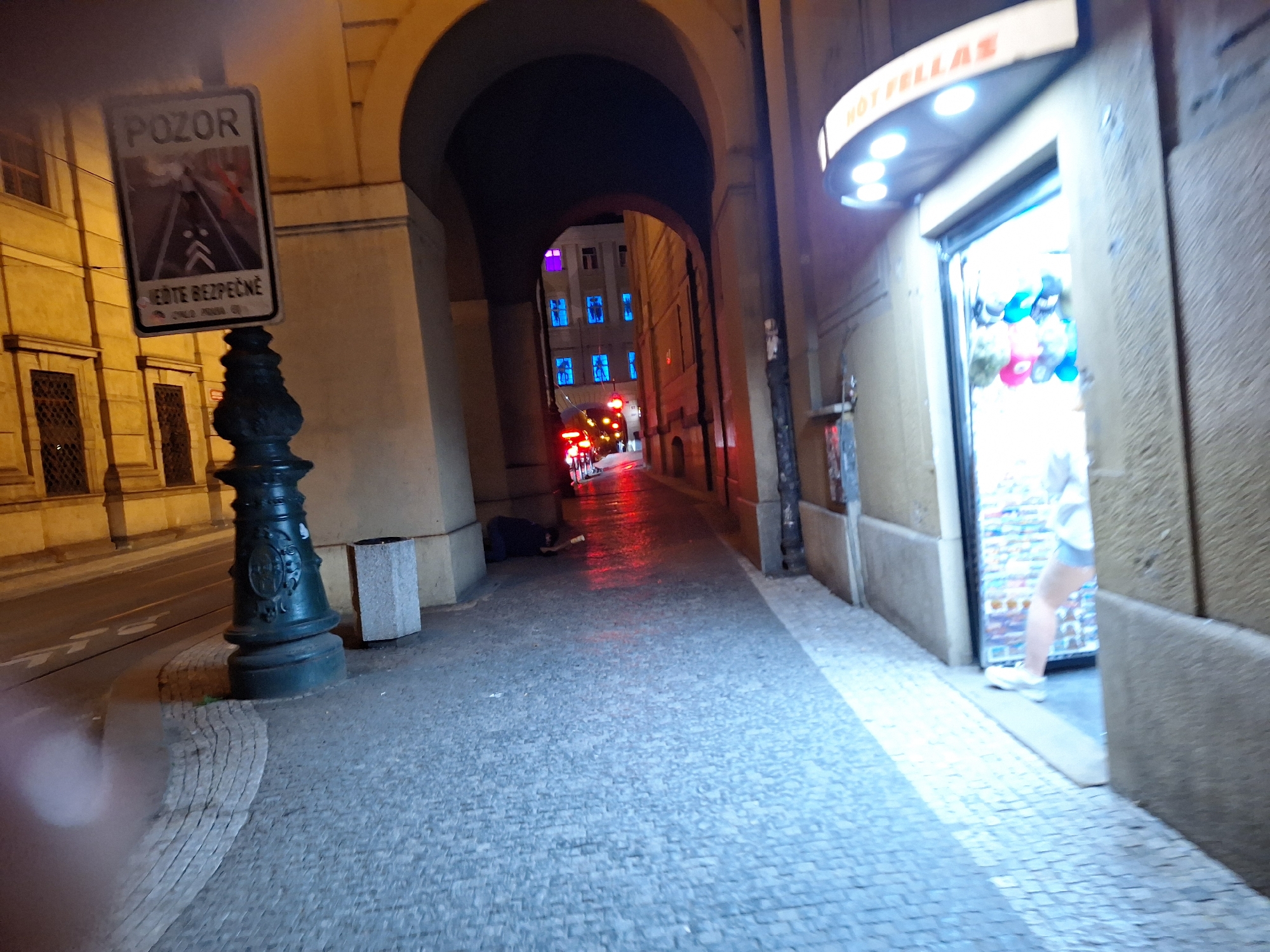 The image depicts a street scene at night in Prague, Czechia. In the foreground, a stone sidewalk curves to the left, paved with cobblestones. A dark green, ornate lamppost stands on the sidewalk, and to its right is a light gray trash receptacle. A street sign with the word "POZOR" (Czech for "CAUTION") and a depiction of a road with arrows is mounted on the lamppost. Further down the street, an arched passageway leads to a narrow street lined with buildings. One building has several windows illuminated with bright blue and purple lights. A person is lying on the ground in the passageway, appearing to be asleep or unwell, with their body partially obscured by darkness. To the right of the passageway, a storefront with a curved awning displays the partially legible text "HOT FELLAS" and is illuminated by several spotlights. Inside the store, colorful merchandise is visible, and a person's legs and lower torso are seen standing near the entrance. Red brake lights from a vehicle are visible in the distance, indicating traffic on the narrow street. The overall atmosphere is dimly lit, with artificial lighting from streetlights, buildings, and the storefront creating pockets of illumination and shadow.