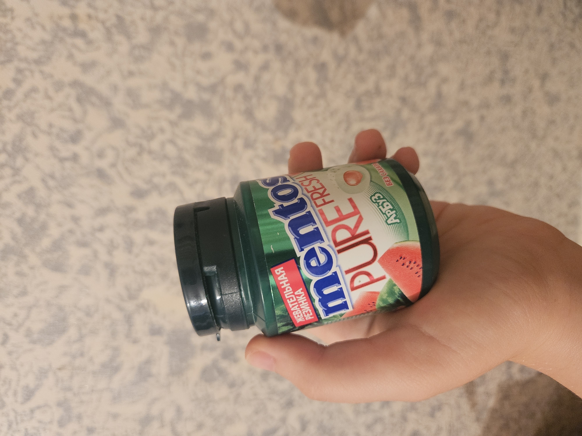 An individual's hand is holding a dark green bottle of Mentos Pure Fresh watermelon flavored chewing gum. The bottle has a dark green cap and a label that features the Mentos logo, the words "PURE FRESH," images of sliced watermelons, and Russian text that translates to "chewing gum," "watermelon," and "sugar-free." The background is a textured, light-colored wall. The image was taken in Unknown, Unknown.