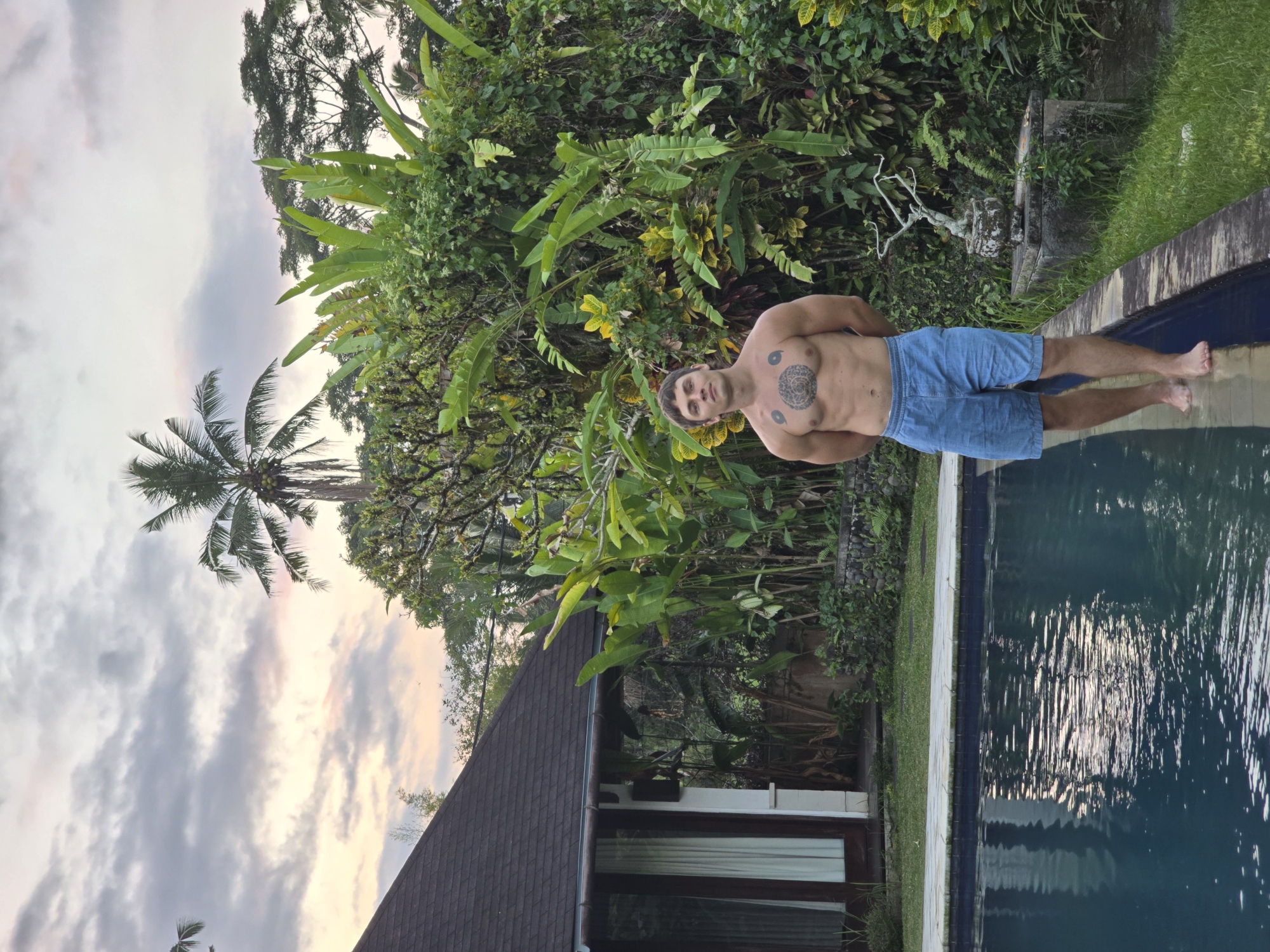 The image, oriented incorrectly and rotated 90 degrees counter-clockwise, depicts a young man standing at the edge of a swimming pool amidst a lush tropical setting. The man, shirtless and wearing light blue shorts, has dark hair and looks directly at the viewer with a neutral expression. He displays several tattoos, including a prominent circular, mandala-like design centered on his chest and two smaller circular designs on his upper arms. His bare feet are partially submerged in the dark blue water of the pool.
The background is dominated by dense tropical vegetation, featuring large-leafed plants, palm trees, and various green foliage. To the left, a section of a building with a dark, sloped tiled roof and a window is visible. Near the pool's edge, among the greenery, there is a stone planter containing a sculptural, bare branch or bonsai-like plant. The sky, visible above the foliage and building, shows soft grey, pink, and pale yellow hues, indicative of either sunrise or sunset. The overall scene suggests a tranquil, outdoor environment, possibly a resort or private residence in a tropical region. No visible text is present in the image.