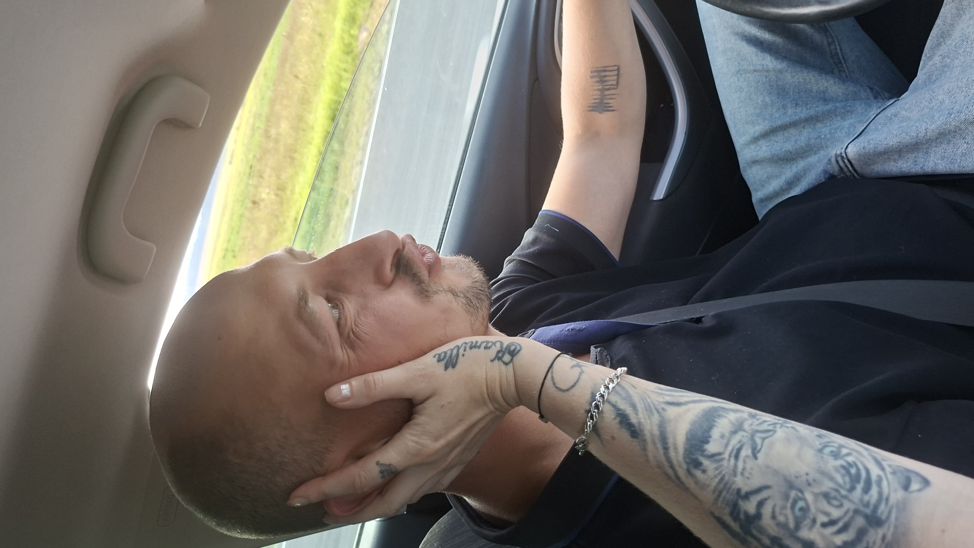 In the image, a man with a shaved head and a goatee is driving a car, looking to the side. A woman's hand is touching his cheek, and her arm, adorned with tattoos, is visible. The tattoos include the name "Camilla," a crescent moon, and a tiger's face. The man is wearing a black shirt and blue jeans. The car's interior is visible, including a seatbelt and a handle on the ceiling. Outside the car window, a rural landscape with green fields and a road is seen. The scene is likely captured while traveling on a road in or around the city of Lyubanskiy Sel'skiy Sovet, Belarus, given the visual cues of a rural Belarusian landscape.