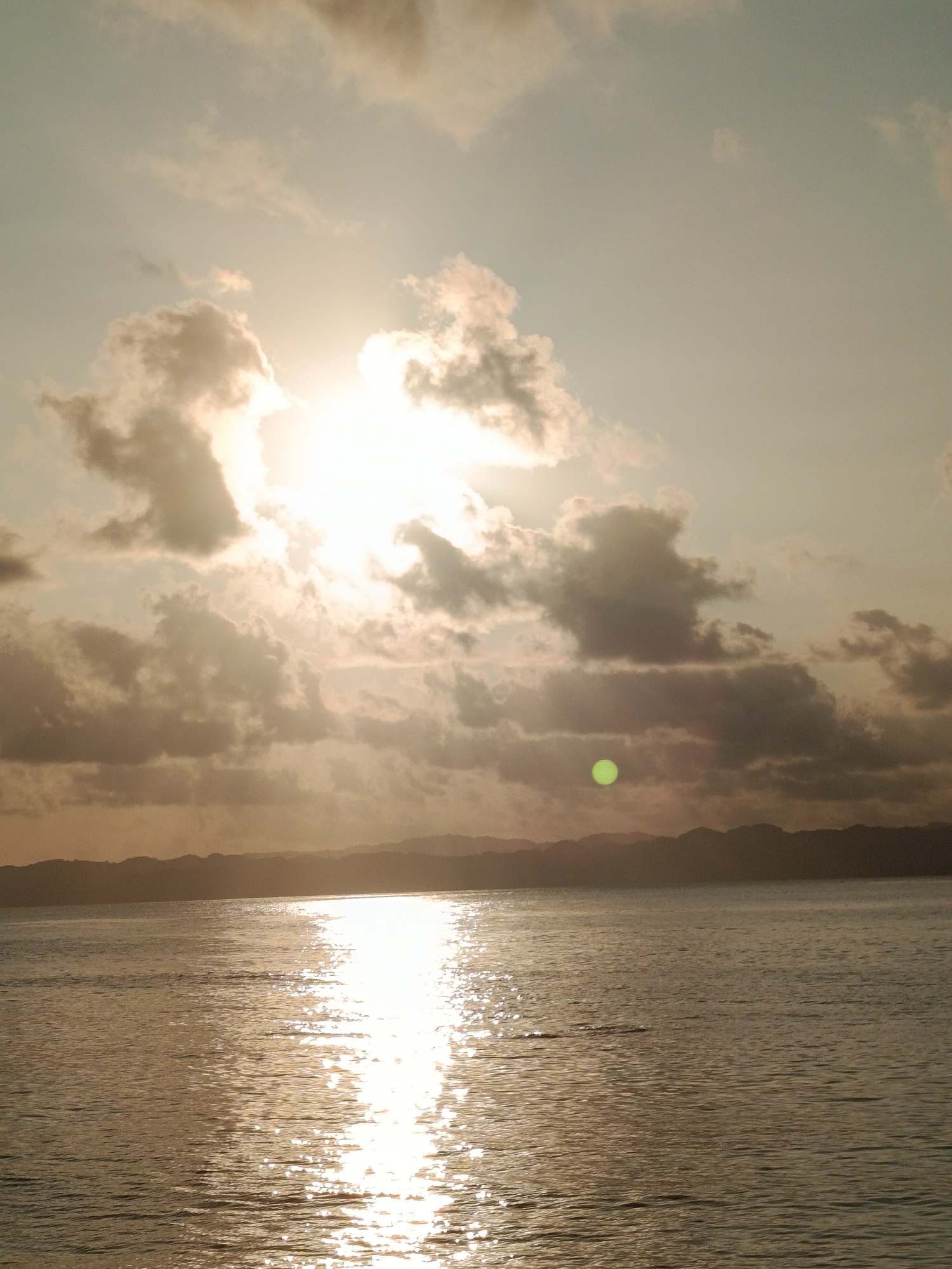 The image captures a serene seascape at sunset. The sun, a bright, diffused orb, is partially obscured by scattered clouds, casting a warm, golden glow across the sky and the calm water. The water reflects the sunlight, creating a shimmering path that leads towards the horizon. In the distance, a silhouetted mountain range forms the backdrop, its contours softened by the fading light. The overall atmosphere is tranquil and peaceful, suggesting a natural landscape rather than an urban environment. There are no people or specific man-made structures visible that would indicate a city location.