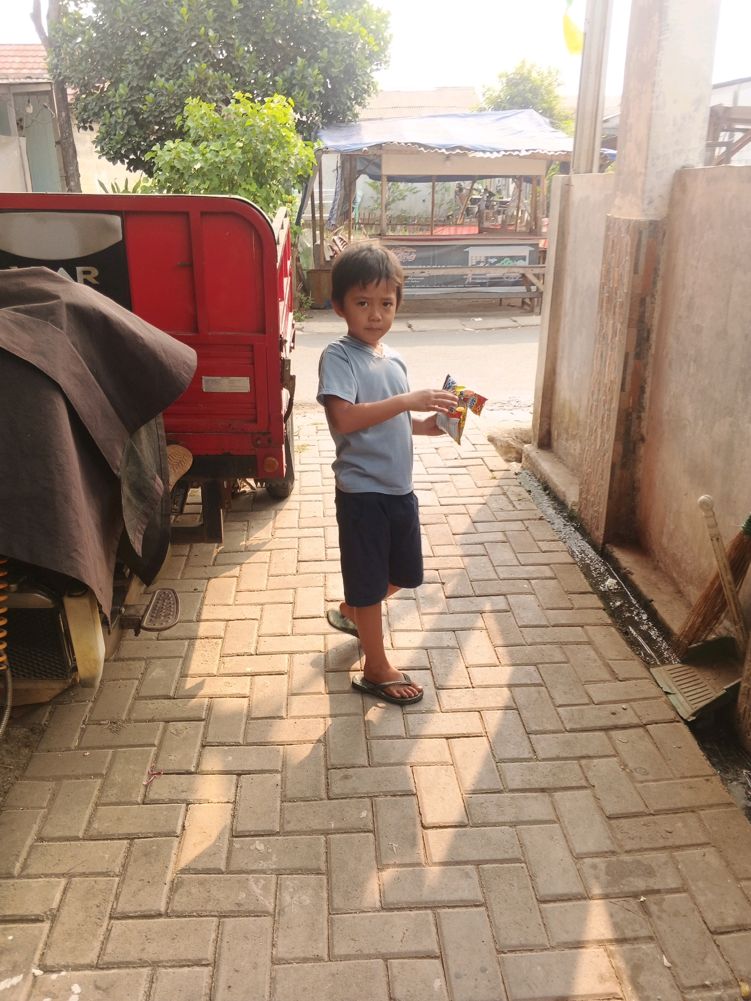A young boy, likely between five and seven years old, stands on a paved walkway in Kadu, Indonesia. He is wearing a light blue t-shirt and dark blue shorts, with black flip-flops on his feet. In his hands, he holds a small, colorful package, possibly snacks, looking towards the camera. The walkway is made of rectangular concrete pavers laid in a herringbone pattern. To his left, a red truck bed is visible, partially covered with a dark tarp. Behind him, a low building with a blue tarp roof, possibly a small shop or stall, is situated across a street. The scene is illuminated by warm, late afternoon sunlight casting long shadows. A concrete wall runs along the right side of the walkway, with brooms and a drain grate leaning against it. The environment appears to be an urban or semi-urban setting with residential buildings and vegetation in the background.
