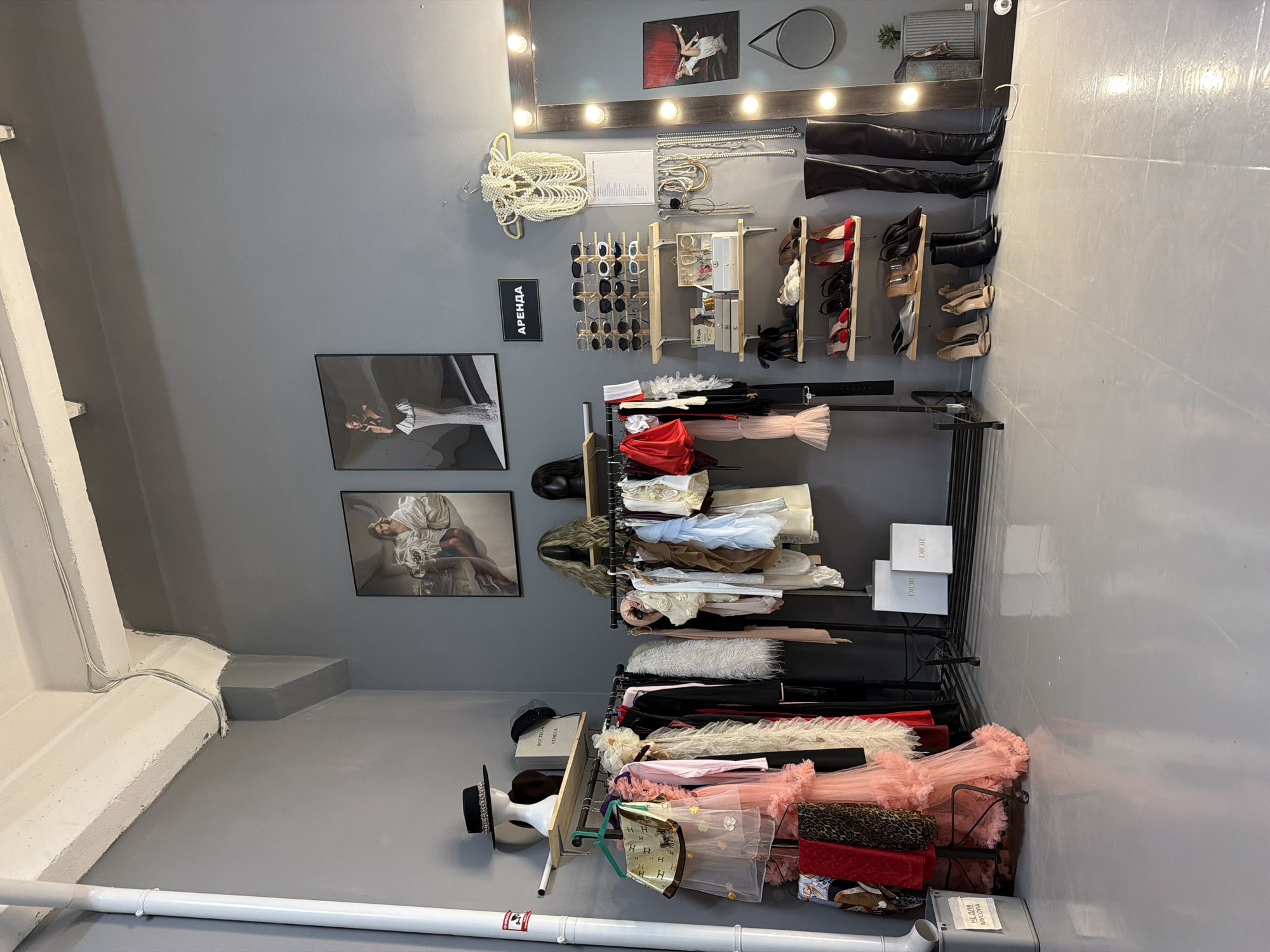 The image depicts an interior space, likely a dressing room or closet, within the city of Kazan, Russia. The walls are painted a neutral gray.
Several items are displayed. On the wall are framed art pieces, a mirror with integrated lights, clothing accessories such as necklaces and sunglasses, and footwear including boots and shoes arranged on shelves. Clothing racks are stacked with apparel, including dresses, faux fur, and bags.
Other details include a radiator pipe running along the floor, and a sign with text written in Russian, "Аренда." The style of the clothing and accessories suggests a high-end retail setting or a well-organized personal wardrobe.