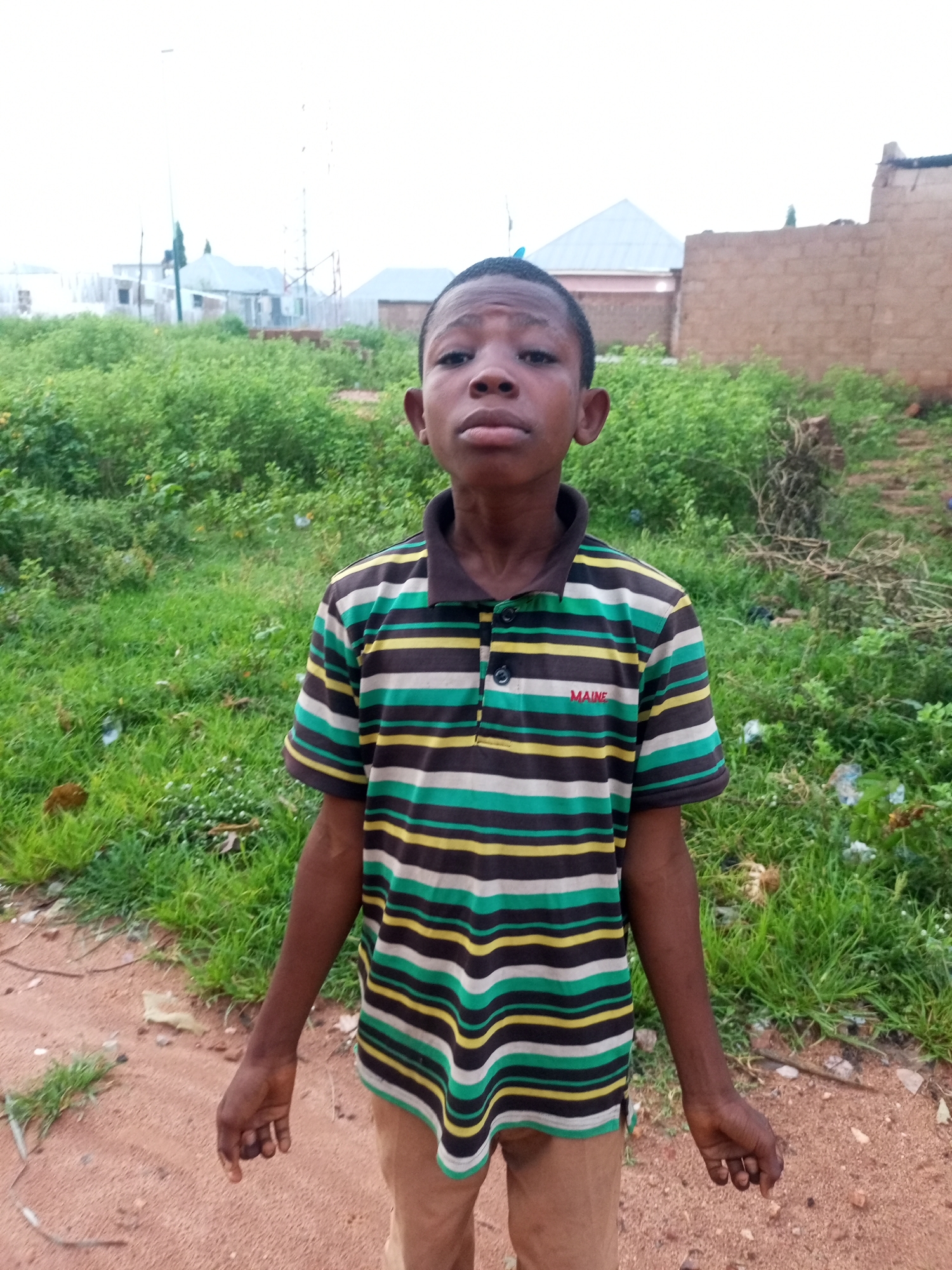 A young boy stands in an outdoor setting, possibly in the city of Tempure, Nigeria. He is wearing a striped polo shirt with black, green, yellow, and white horizontal bands, and beige pants. His shirt has a dark collar and a small red label with the word "MAINE" written on it. The background consists of a patch of green vegetation, some loose soil, and in the distance, buildings and what appear to be makeshift structures, suggesting a semi-urban or rural environment. The sky is overcast. The boy is looking directly at the camera with a neutral expression.