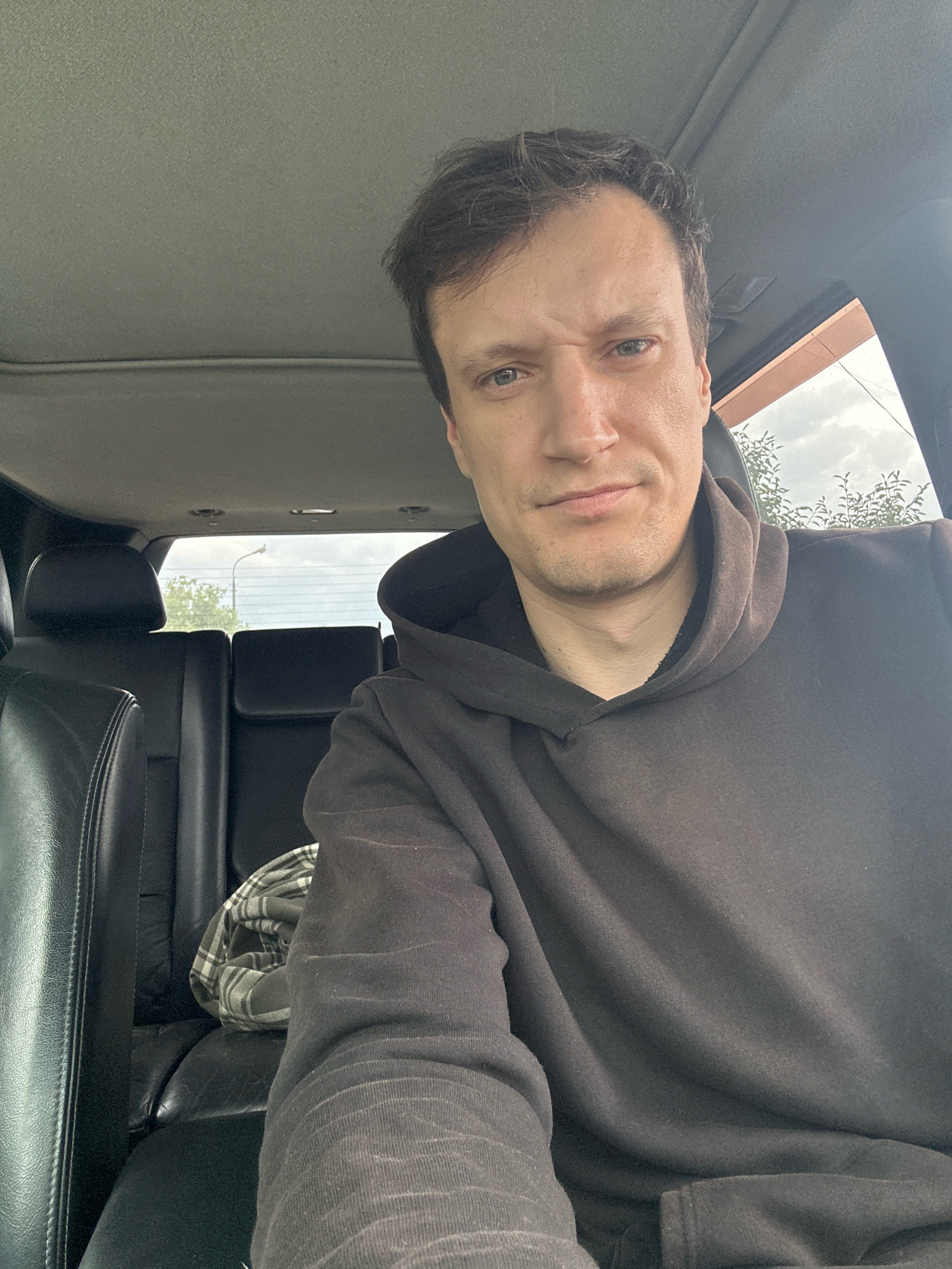 A man is sitting in the passenger seat of a vehicle, looking towards the camera with a slightly furrowed brow. He is wearing a dark hooded sweatshirt. In the background, the back seats of the car are visible, along with a patterned blanket folded on the seat. Through the car windows, a cloudy sky and trees can be seen. The image appears to be taken during the daytime, and the location is Makiv, Ukraine.