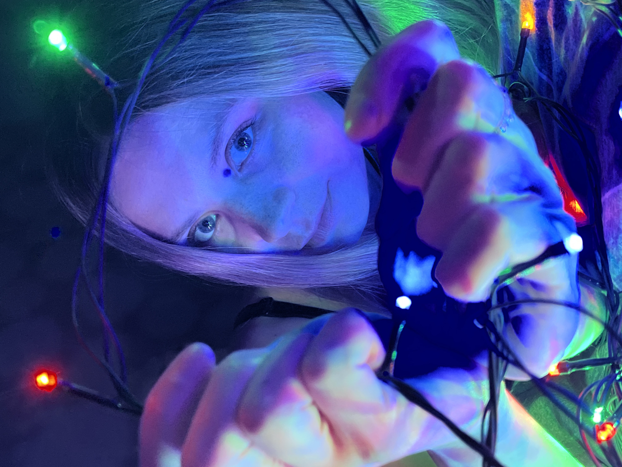 A low-angle close-up photograph shows a fair-skinned person, presumably female, with long, light-colored hair, illuminated by multicolored Christmas lights. The person has blue eyes and a prominent mole on their face. Their hands are visible in the foreground, entangled with the strands of Christmas lights. The lights emit a spectrum of colors, including red, green, orange, and blue. The background is dark, suggesting an indoor setting. The image shows the person's interaction with the Christmas lights. There is no other indication of location in the city of Chaikovsky, Russia, or further forensic details.