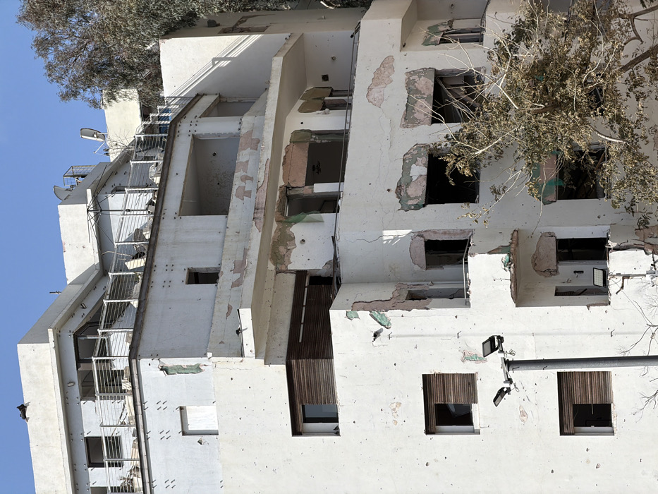 A multi-story residential building in Tel Aviv, Israel, is depicted under a clear sky. The building exhibits extensive damage consistent with projectile impacts. The white exterior walls show numerous small, circular indentations and larger areas of spalling, where plaster has peeled away, exposing the underlying concrete and hints of green and pink substrate. Cracks are visible in the concrete walls. Multiple windows on various levels are either missing glass panes or have damaged wooden shutters. Balconies on several floors appear compromised, with railings bent or partially missing, and debris scattered on their surfaces. The upper levels feature open terraces with metal railings; one terrace holds a partially collapsed outdoor umbrella and what appear to be two satellite dishes. A tree with sparsely leaved branches stands in the foreground, partially obscuring the lower levels of the building. A dark gray street light pole with two rectangular light fixtures is visible on the lower left. No individuals are present in the frame.
