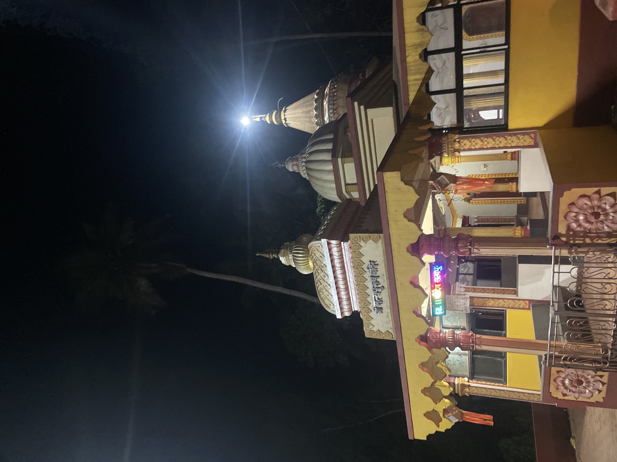 The image shows a Hindu temple at night in Redi, India. The temple is brightly lit with a spotlight illuminating its spires. The building has multiple tiers and levels, decorated with ornate carvings and colorful paint. The temple’s facade includes an entrance with columns and intricate floral patterns. A sign in Hindi script is visible on one side. A multi-story section with windows and a balcony is attached to the temple. Tall palm trees are faintly visible in the background, blending into the dark night sky. A wrought iron fence surrounds a part of the building.