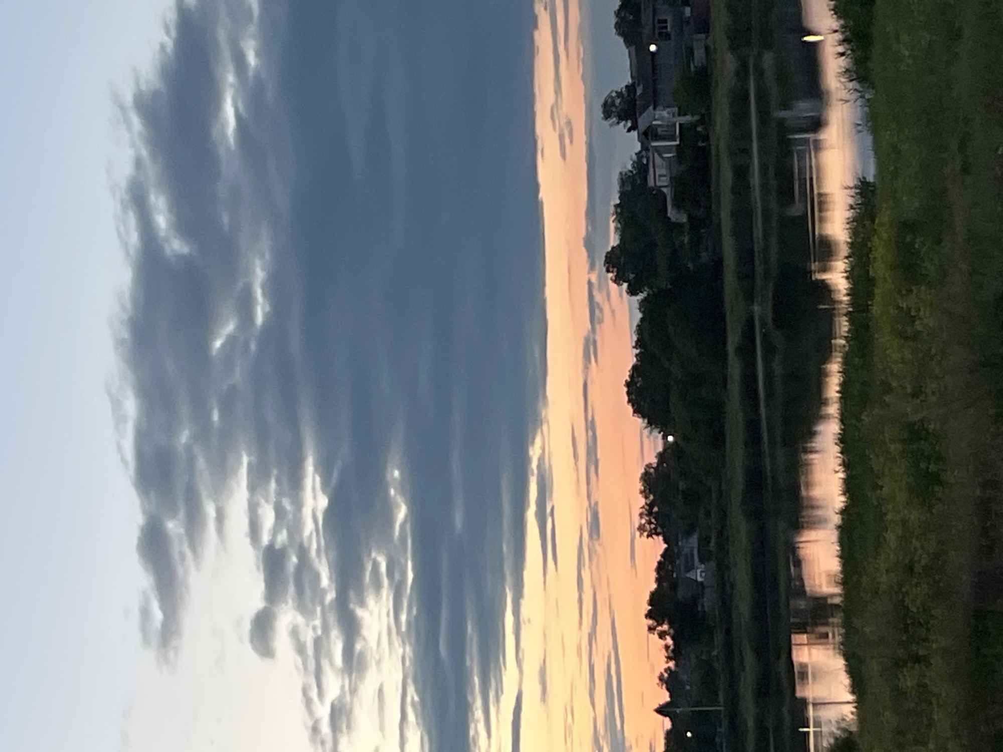 The image captures a twilight or dawn scene beside a body of water. The sky is divided into a blue area with textured clouds on the left and a pinkish-orange section on the right, suggesting sunrise or sunset. A line of trees and buildings runs along the right edge of the frame, with their reflections visible in the water. Grass or vegetation borders the water on the right side. The presence of houses and streetlights near the water could suggest that the location is near a residential area of a city. No people are clearly visible. Given that Kryvyi Rih is mentioned as a location, the waterway could be a part of the Saksahan River or any of the other smaller water bodies in the city. Specifics like the exact type of architecture cannot be determined without a sharper focus, and the scene offers an atmospheric depiction of dawn or dusk.