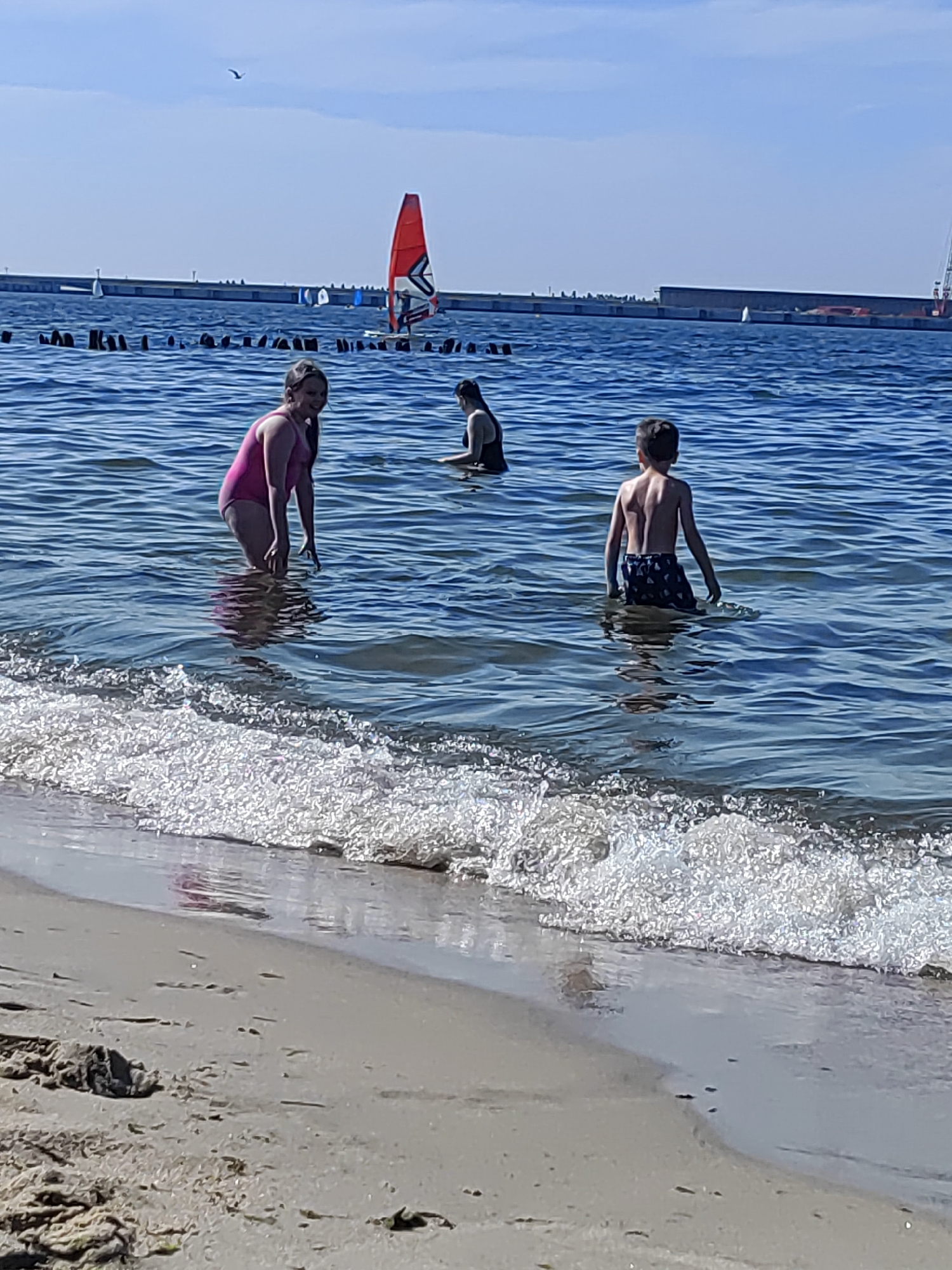In Pionersky, Russia, on a sunny day, three young people are enjoying the water at the beach. A girl in a pink swimsuit stands in the shallow water, facing the camera and smiling. To her right, a person wearing a black swimsuit is partially submerged in the water. Further to the right, a boy in patterned swim trunks stands with his back to the camera, looking out at the sea. Gentle waves are breaking on the sandy shore in the foreground. In the background, a breakwater extends into the sea, with several sailboats and a red windsurfing sail visible. The sky is clear blue.