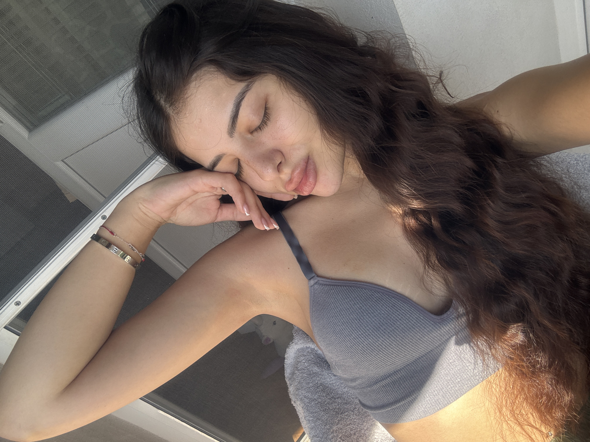 A young woman with long, dark, wavy hair is lying down, her eyes closed, and her head resting on her hand. She is wearing a grey ribbed bra. Her left arm is visible, adorned with two bracelets. A grey towel is partially visible near her chest. A window with a screen is in the upper left corner. The scene appears to be indoors, possibly in Plovdiv, Bulgaria, although specific city landmarks are not visible in the image.