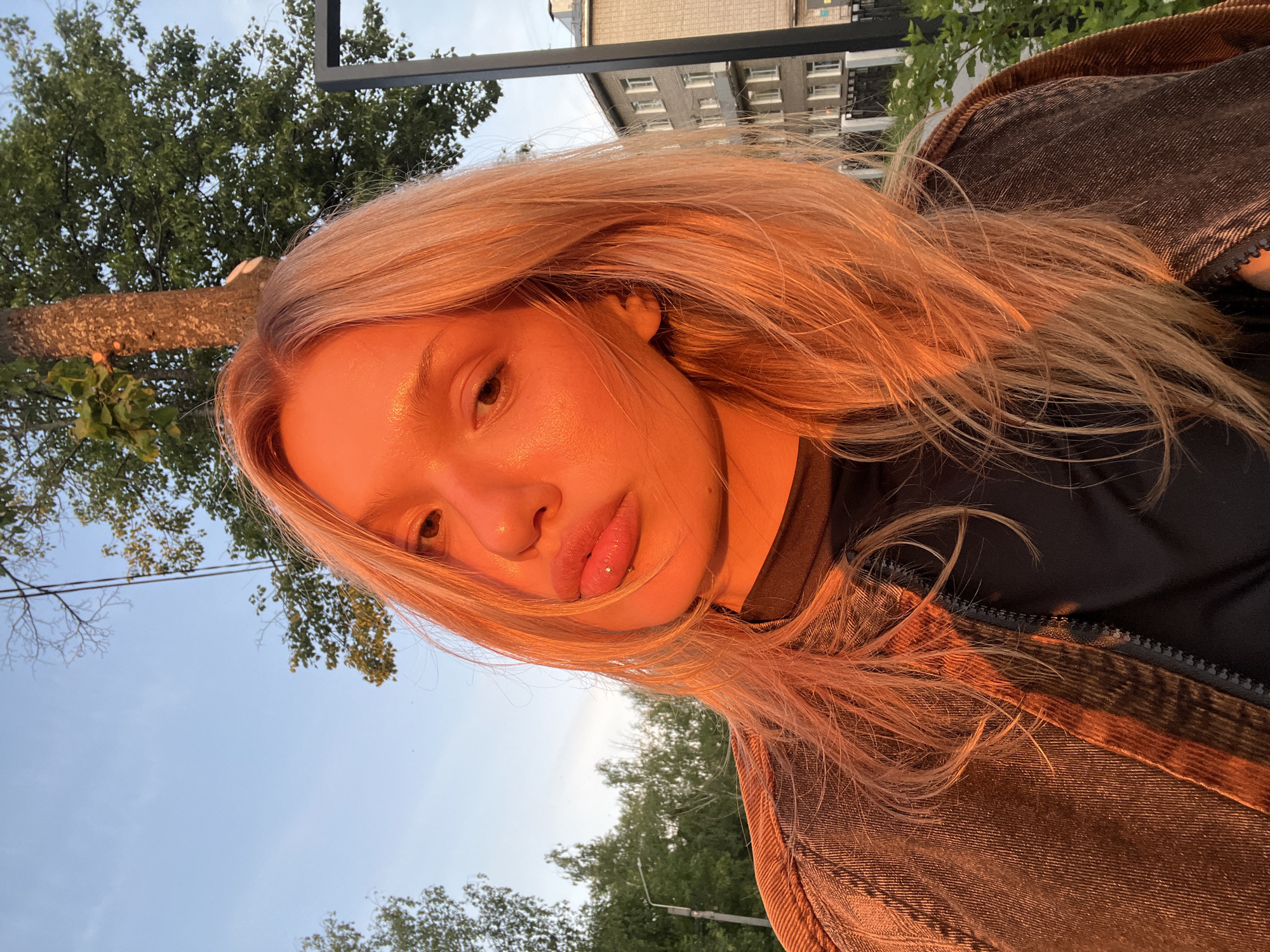 A portrait-style image features a young woman outdoors, captured during what appears to be the golden hour of sunset or sunrise, evidenced by the intense warm, golden-orange light illuminating her. The photo is taken at a tilted, dynamic angle, with the woman's head angled downwards and to her right, occupying the left and center of the frame.
The subject has long, light blonde hair that glows under the warm light. Her face is partially visible, characterized by full lips with a small, sparkly stud piercing on her lower lip. Her eyes, appearing brown or dark, are directed towards the viewer. Her skin reflects the golden light, giving it a luminous, slightly dewy appearance.
She is dressed in a dark-colored top, possibly black, underneath a jacket made of a textured, reddish-brown or brown fabric, resembling corduroy or heavy denim, with a visible zipper.
The background includes both natural and urban elements. A large tree trunk and dense green foliage are visible to the left. Above and behind the woman, a clear, light blue sky with some faint clouds stretches across the upper part of the frame. In the upper right, a multi-story building with multiple windows, characteristic of an apartment block or urban structure, is discernible. The setting suggests an outdoor urban or suburban environment, consistent with Chaikovsky, Russia. There is no visible text in the image. The prominent lighting creates a warm and intimate atmosphere.