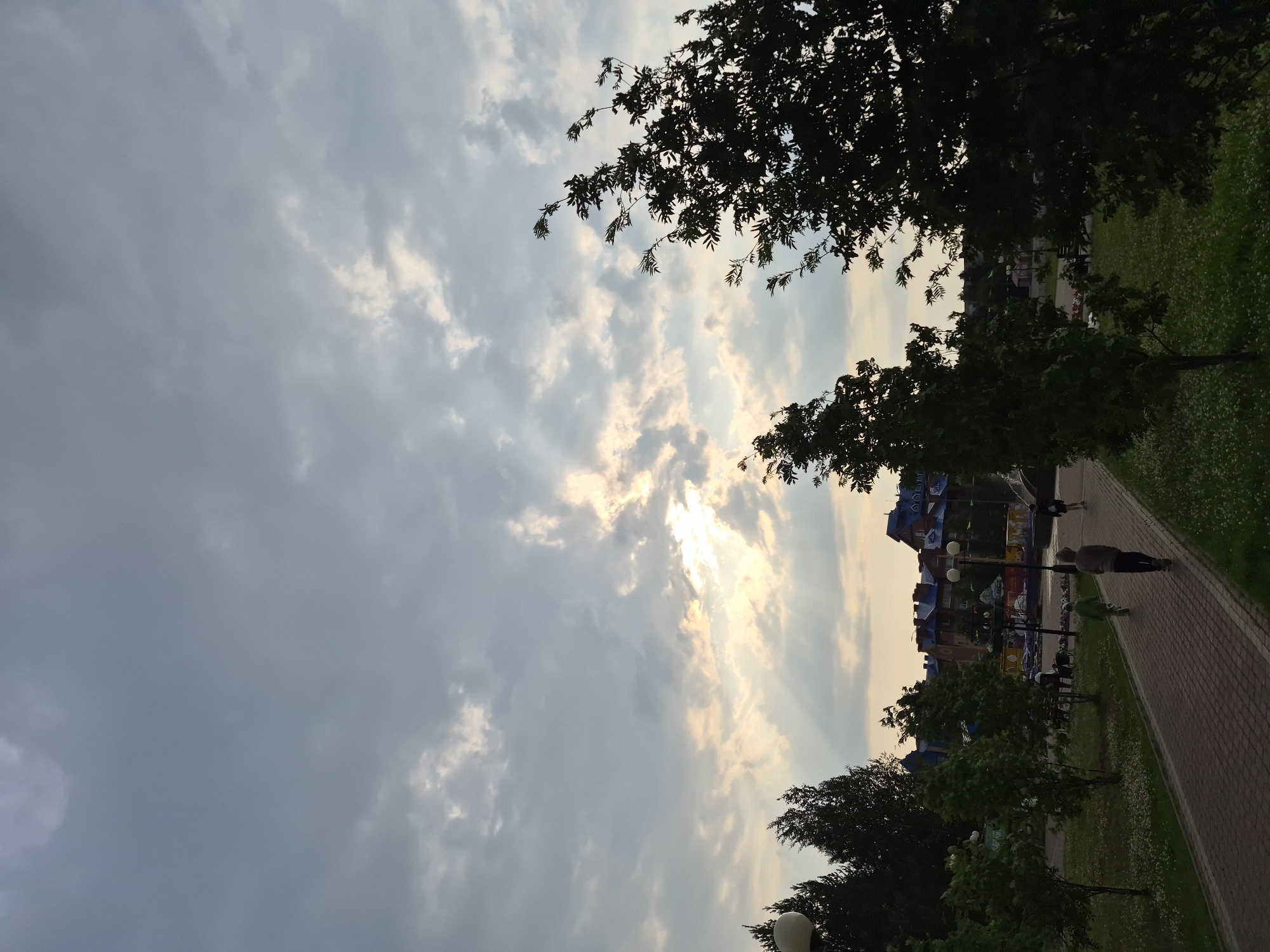 The image depicts a park scene in Nyagan, Russia, during what appears to be late afternoon or early evening, given the soft light filtering through the clouds. The sky is dramatic, with rays of sunlight piercing through scattered clouds, creating a dramatic and somewhat moody atmosphere. The sun itself is partially obscured, suggesting it's low in the sky.
In the foreground, silhouetted trees frame the scene on the left and right. The trees on the right have dark, leafy canopies. A paved walkway, made of reddish-brown bricks, runs along the right side of the image, bordered by a lush green lawn dotted with small white flowers.
Along the walkway, several people are visible. One person in a light-colored top and dark pants walks away from the viewer. Further ahead, another person in a green top walks in the same direction. In the distance, more figures are seen walking, suggesting a leisurely stroll.
Behind the walkway and lawn, a row of buildings with distinctive blue roofs is visible. These buildings have a somewhat whimsical, storybook architectural style. There are also streetlights and what appear to be playground elements or amusement rides with colorful structures.
The overall impression is one of a peaceful, urban park at the end of the day, with people enjoying the open space as the sun begins to set. The weather seems fair, with the clouds adding a visually interesting element to the sky. No visible text is present in the image.